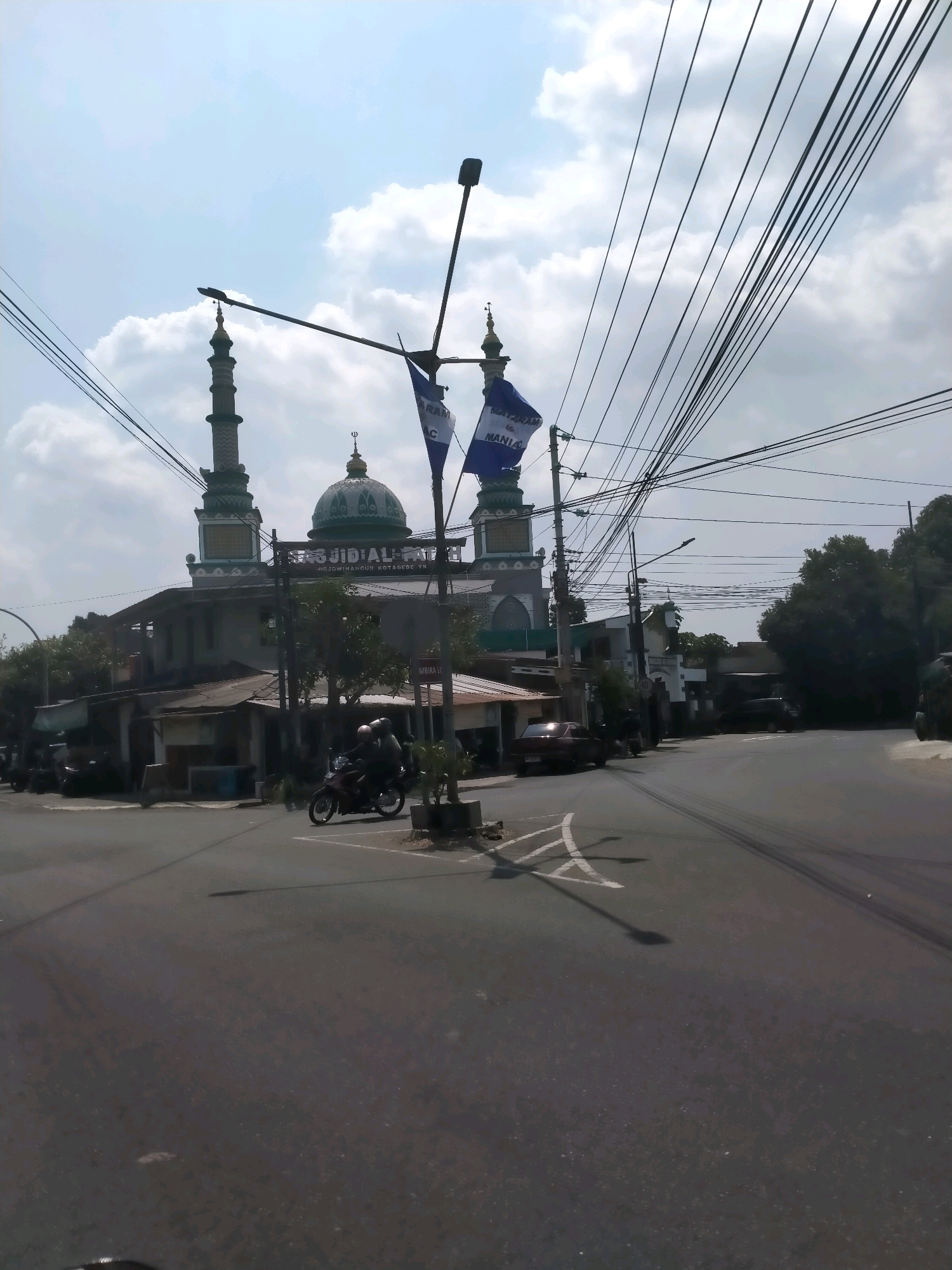 In this image, a mosque with a green dome and two minarets is visible, identified by signage as "Masjid Al-Fatah" in Yogyakarta, Indonesia. A motorcycle with two occupants is in motion on the road in front of the mosque. The scene is characterized by a paved road, several buildings, and a sky with clouds. Overhead, numerous electrical wires crisscross the frame, and a streetlamp is present. The overall impression is a snapshot of a typical day in a city environment.
