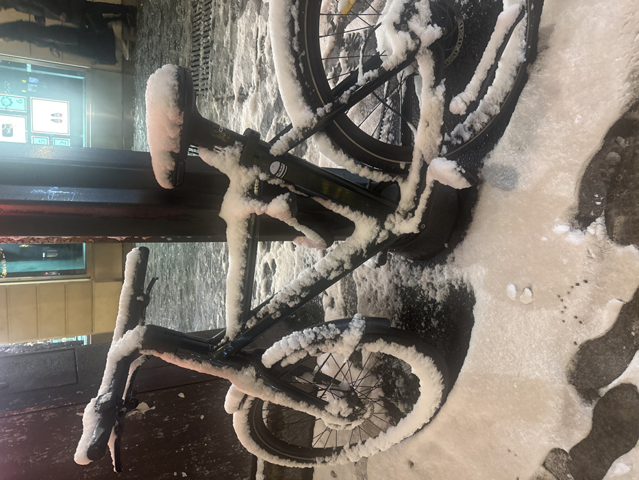 A black electric bicycle, heavily covered in snow, leans against a dark metallic structure outdoors. Snow also blankets the surrounding pavement. The bicycle's frame, handlebars, wheels, and seat are all adorned with a thick layer of snow. Reflections from nearby lights are visible on the dark structure and the bicycle. A blurred storefront window with illuminated displays appears in the background.