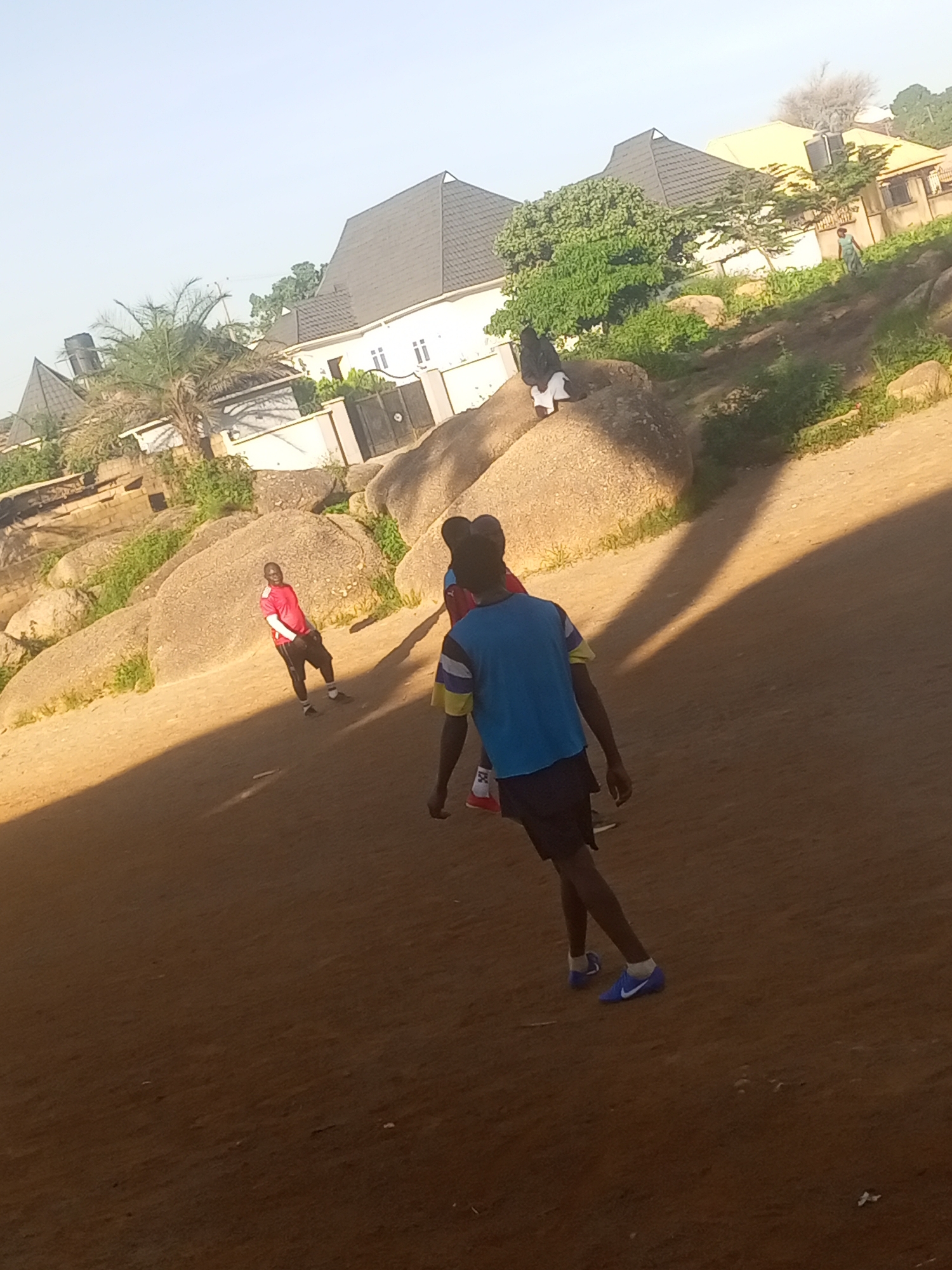 In Jos, Nigeria, a group of young men are observed in an open area, possibly a field or a clearing, with large rock formations and residential buildings in the background. One individual, wearing a red t-shirt and black shorts, stands facing the camera, appearing to be mid-stride. Another person, seen from behind, is wearing a blue and yellow striped shirt and dark shorts, with blue athletic shoes. A third individual, also seen from behind, is wearing a white garment with black accents, sitting on a large rock. The ground is composed of dirt and sand, with sparse vegetation and shadows cast by the sun. In the background, the roofs of houses and some trees are visible, suggesting an urban or semi-urban environment. The overall scene depicts a casual outdoor activity involving young men in a rocky, residential setting in Jos.