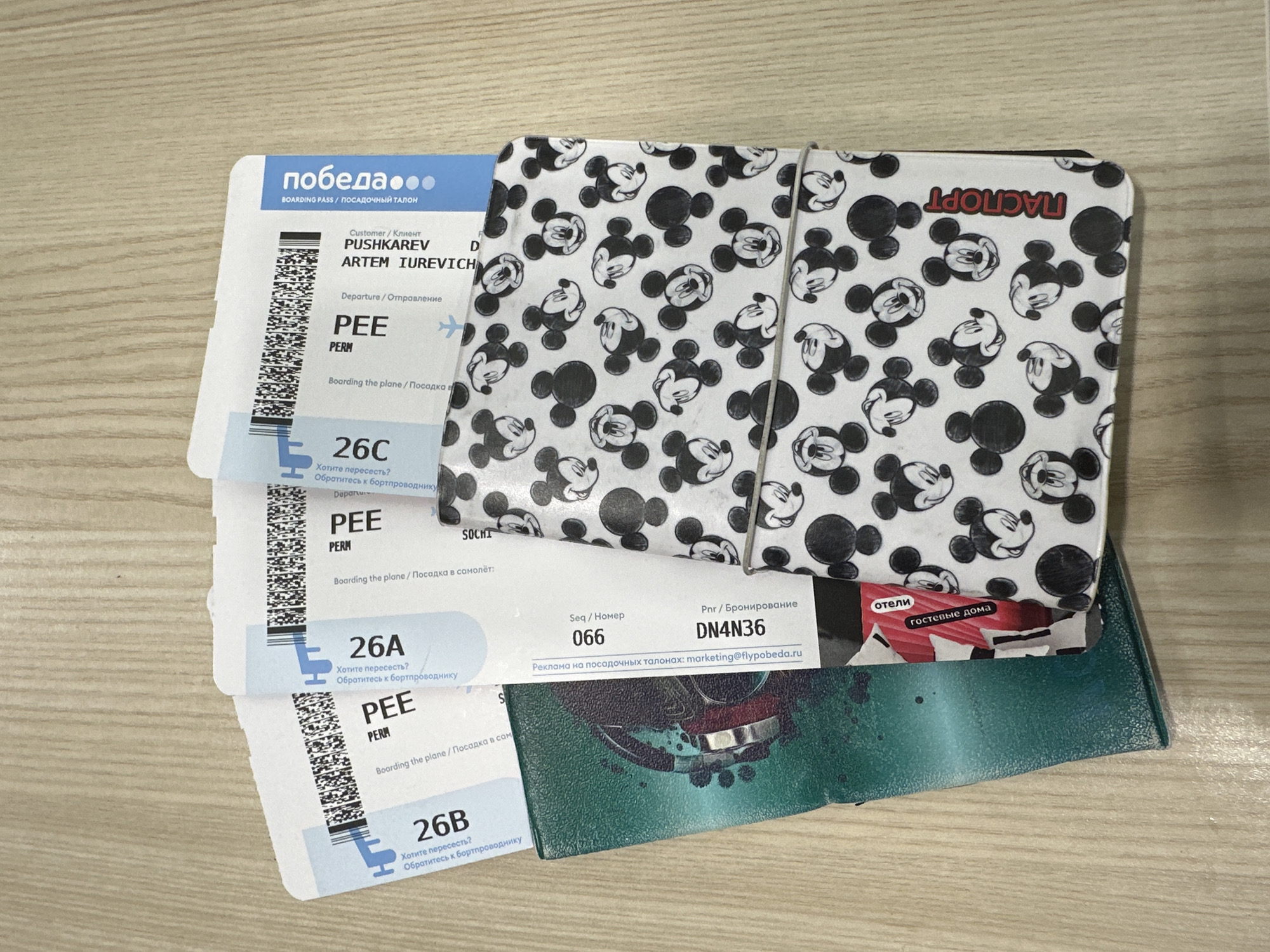 The image shows three Pobeda boarding passes stacked on top of a Mickey Mouse passport holder and a green wallet, all placed on a light-colored wooden surface. The boarding passes are for the same departure location, PEE (Perm), and have seat numbers 26C, 26A, and 26B. The customer name on the passes is PUSHKAREV ARTEM IUREVICH. The passport holder has a white background with a black Mickey Mouse pattern and the word "ПАСПОРТ" printed on it in red. The phone holder has a graphic of a person's face and red, blue, and green colors. The image contains no people or actions. There is no information about Boldino, Russia, in the image.