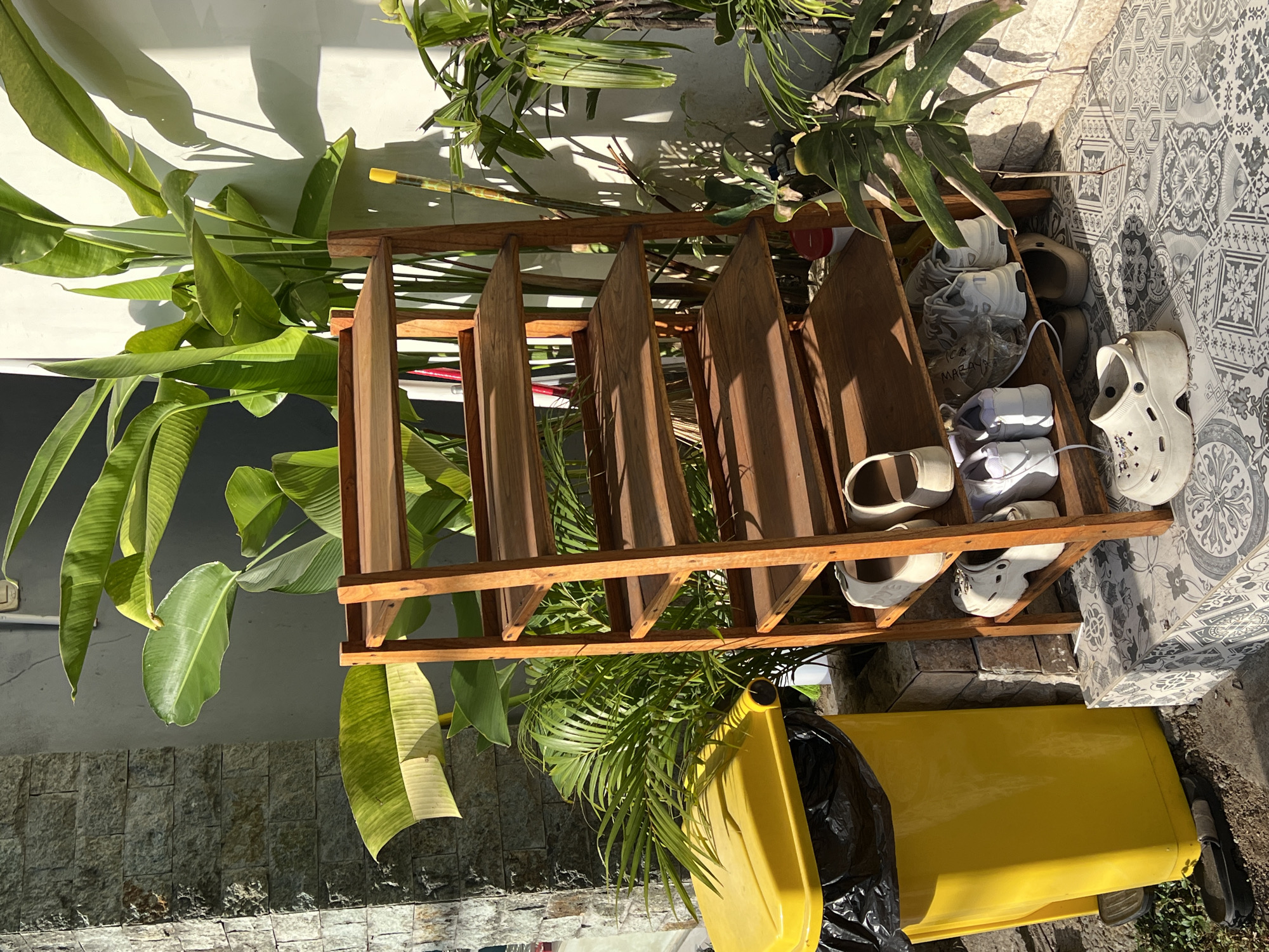 The image shows a shoe rack with shoes in it, placed outside on a patterned tile floor. The shoe rack is made of brown wood. Shoes of various colors, including white, beige, and gray, are visible inside the rack. Lush green plants surround the shoe rack, and a yellow trash can with a black bag inside is located nearby. The photograph was taken in the city of Ubud District, Indonesia.