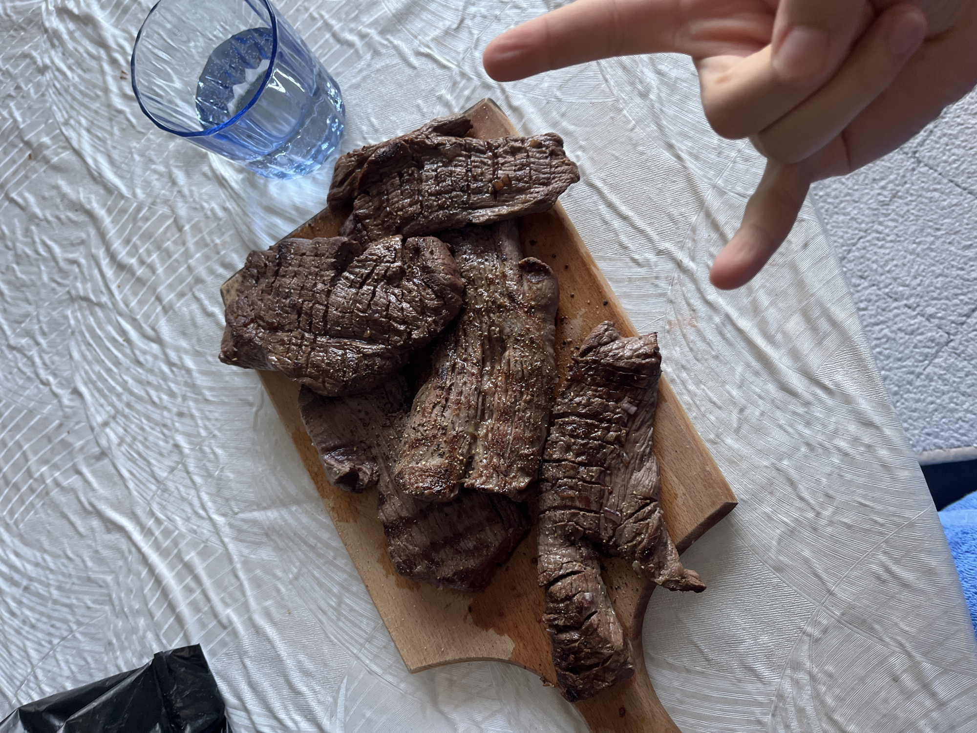 The image, taken from an elevated perspective, displays a meal arranged on a white, textured tablecloth. Central to the composition is a light brown wooden cutting board holding multiple pieces of cooked meat, likely steak, exhibiting grill marks and seasoning. To the upper right of the cutting board, a translucent blue glass filled with water is present. In the upper left corner, a crumpled black plastic bag is partially visible. A human hand, positioned in the lower right foreground, is performing a gesture with the thumb, index finger, and pinky extended, resembling the "I Love You" sign in American Sign Language. The lower edge of the image reveals a partial view of a blue towel or fabric and a grey, textured surface, possibly a floor or rug. The lighting suggests an indoor setting.