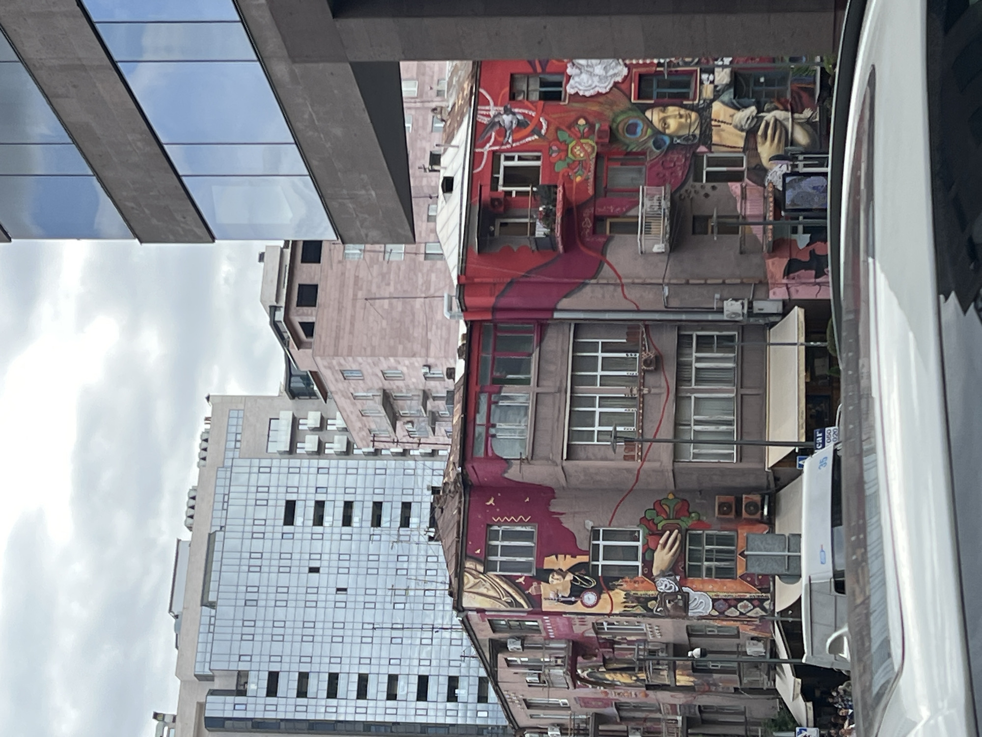 The image shows a street scene in Yerevan, Armenia, with buildings displaying vibrant murals alongside modern architecture. A multi-story building covered in colorful murals, including depictions of people and abstract designs, stands next to a more plain, light pink building. In the background, a taller building with a grid-like pattern of windows is visible, partly obscured by clouds. An adjacent building with glass panels reflecting the sky. A white vehicle on the right side of the image further characterizes the busy street setting.