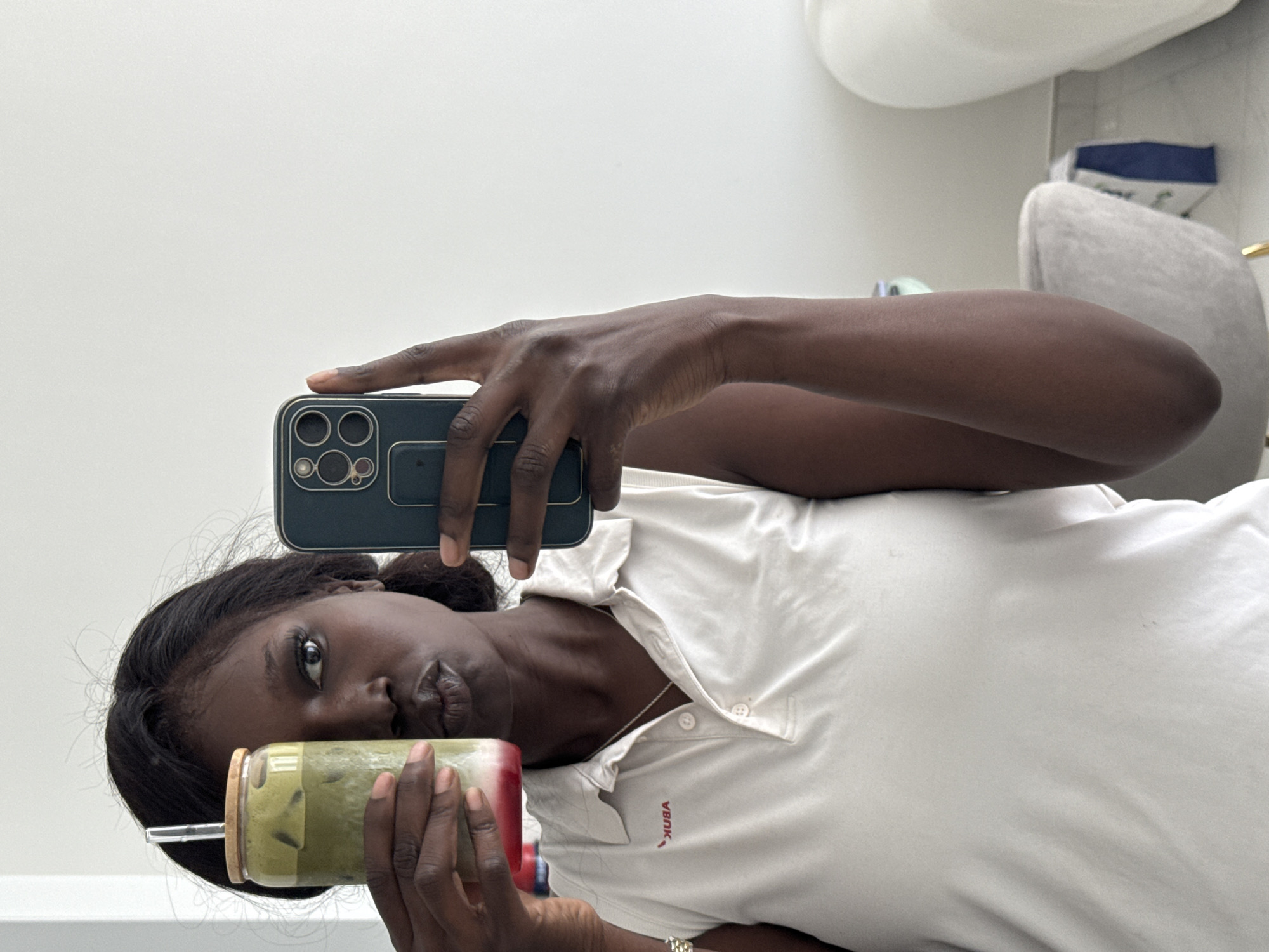 A dark-skinned woman is seen in a selfie, holding a beverage in a glass with a straw in one hand and her phone in the other. She is wearing a white polo shirt with what appears to be a silver necklace and a silver bracelet on her wrist. The phone is in a dark case with multiple camera lenses visible. The background is predominantly white and appears to be indoors with a hint of a chair and other objects in the background. There is no indication of the city of Dubai, United Arab Emirates, or any other specific location in the scene.