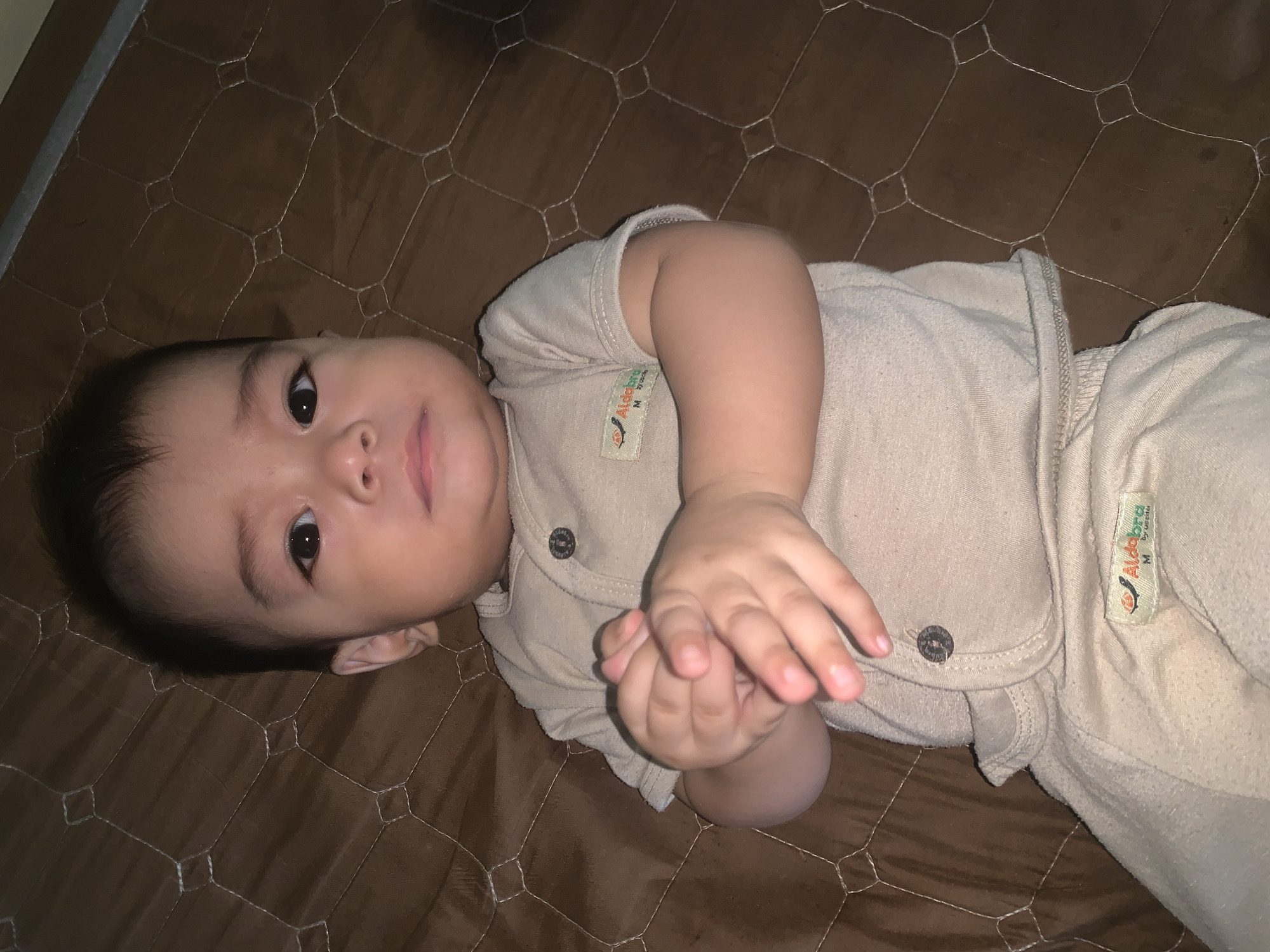 A close-up, eye-level shot captures a baby lying on a brown, diamond-patterned mat, possibly a crib or play mat, in Sungai Meriam, Indonesia. The baby, appearing to be around 6-12 months old, has dark hair and is dressed in a light beige, short-sleeved onesie with two buttons at the neckline and a small, orange and green tag on the left chest. The onesie has a label on the left side of the chest with text that appears to read "Aldo bra M by usita". The baby's hands are clasped together in front of their chest. The background is the brown mat, with a faint shadow on the right side of the frame indicating a light source. The focus is sharp on the baby's face and hands, with a slight blur on the edges.