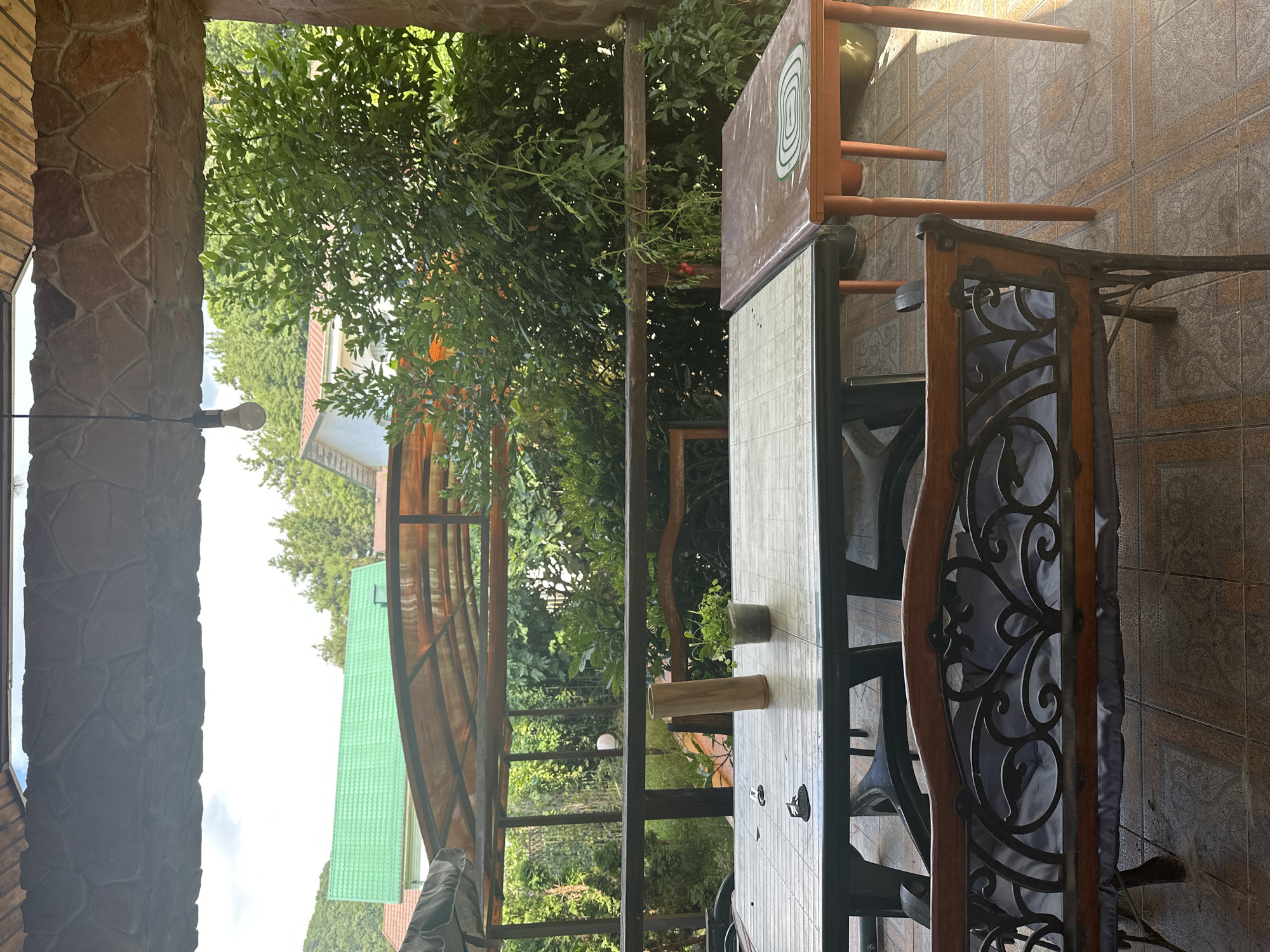 This is a photograph taken outdoors on a sunny day, likely on a porch or patio with a stone-clad overhang. The scene depicts a relaxed outdoor seating area with two tables and a bench. In the foreground, a wooden and wrought-iron bench with a grey cushion is positioned on a tiled floor. Behind the bench, a rectangular plastic table with a light-colored checkered pattern is set up, with dark chairs visible underneath. On this table, there is a tall, cylindrical wooden object, a small potted plant, and two small black spiral-shaped items, possibly mosquito coils. To the right of this table, a smaller, orange-legged table with a brown surface covered by a plastic sheet is visible. The background reveals a lush green garden with various trees and foliage. Beyond the garden, several houses with tiled roofs, one distinctly green, are nestled amongst the trees, and a hillside is visible in the distance. The overall impression is a tranquil setting, potentially a cafe or a private residence's outdoor entertainment space in a suburban or semi-rural area.