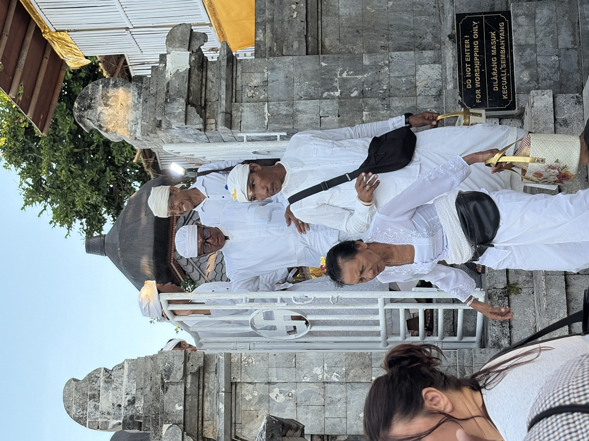 In Kuta Selatan, Indonesia, a group of people dressed in white attire, including head coverings, are descending stone stairs next to a gray stone structure and a white metal gate. Some individuals carry baskets and bags. A sign on the wall reads "DO NOT ENTER! FOR WORSHIPPING ONLY" and "DILARANG MASUK KECUALI SEMBAHYANG." The environment appears to be an outdoor temple or religious site, with trees and a clear sky visible in the background.