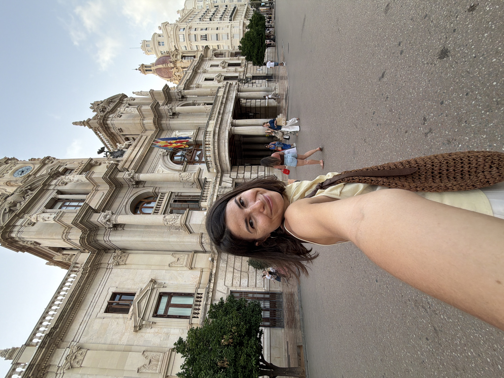 A woman in a yellow top and white skirt is taking a selfie in front of a large, ornate building. She has dark hair and is smiling at the camera. Behind her, several other people are walking on a paved street. The building appears to be made of stone and has classical architectural elements. The sky is cloudy. The scene is set in a city square.