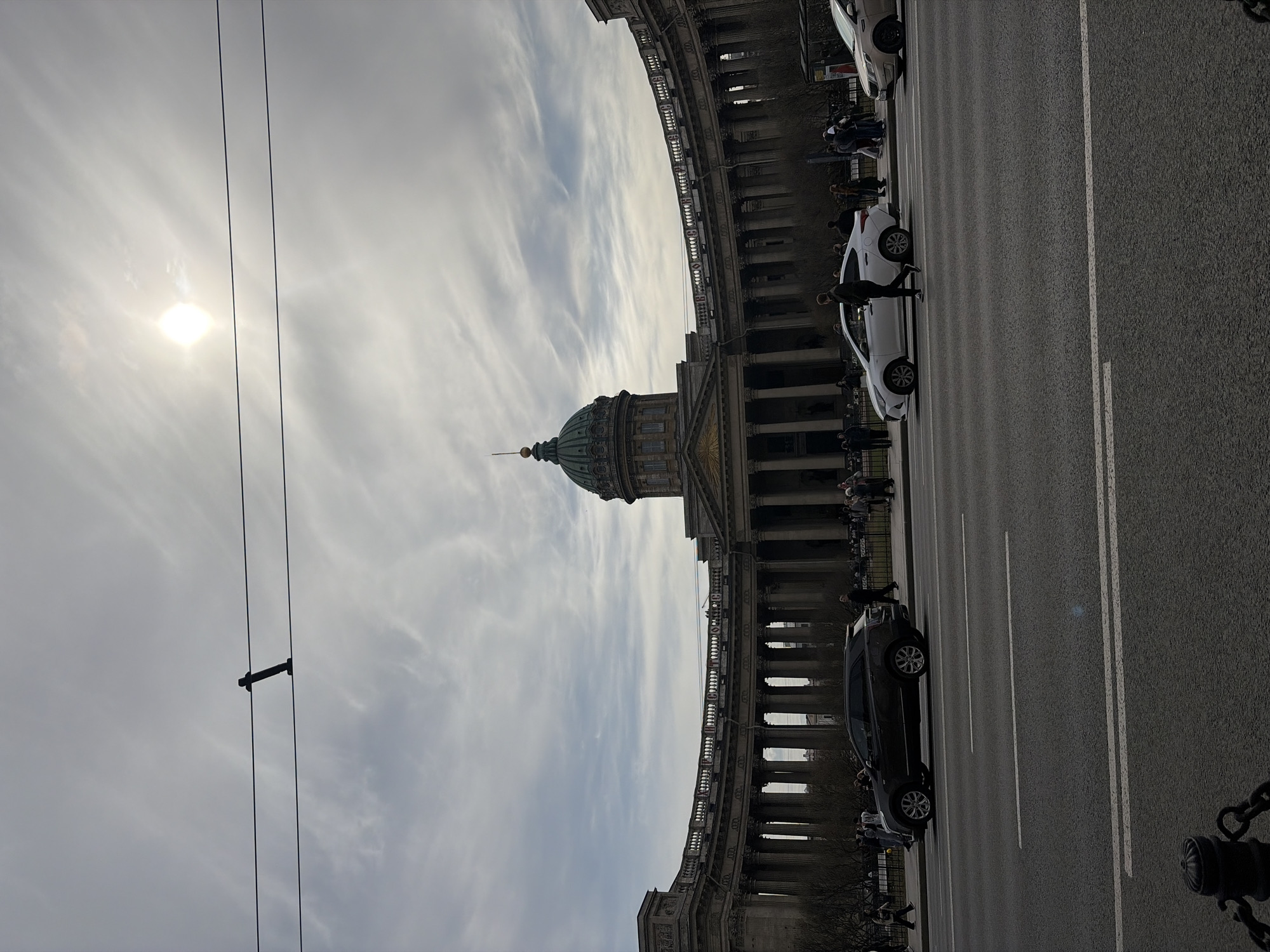 The image depicts a street scene in Saint Petersburg, Russia, featuring the Kazan Cathedral prominently in the background. The cathedral's dome is visible against a cloudy sky with a bright sun to the left. Power lines are visible in the sky. The street has several cars parked or moving along it, and pedestrians are present near the cathedral. A black chain is visible in the lower right corner.