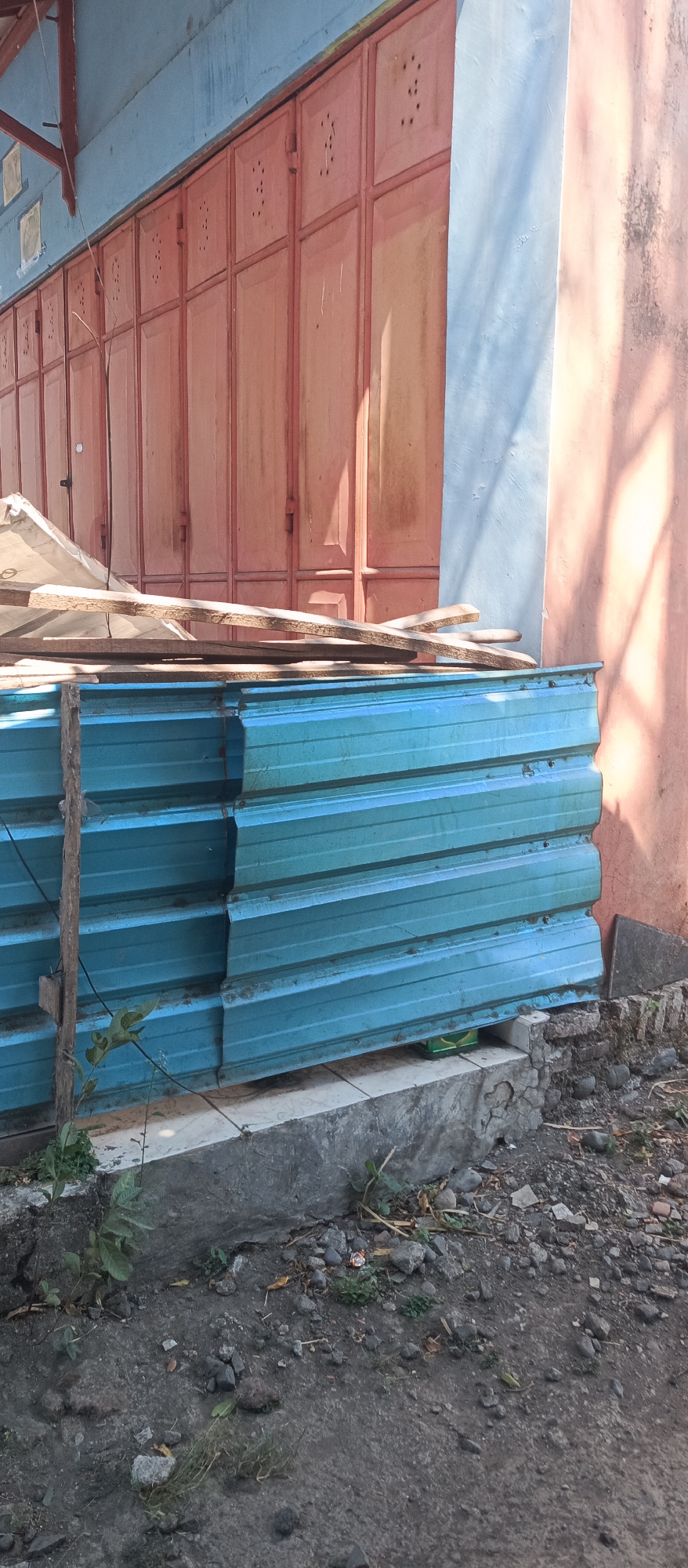 A stack of blue corrugated metal sheets is leaning against a concrete step in front of a building. The metal sheets are bound together with a thin rope and a wooden stick. Above the metal sheets, there are some wooden planks placed on top of a white tarp. The building has a light blue facade and orange metal doors with decorative perforations. To the right of the building, there is a pink wall with shadows cast by tree branches. The ground in front of the building is covered with dirt, small rocks, and patches of grass. There is also a visible concrete curb and a small green plant growing near the metal sheets. The scene appears to be an outdoor storage area next to a commercial or residential building.