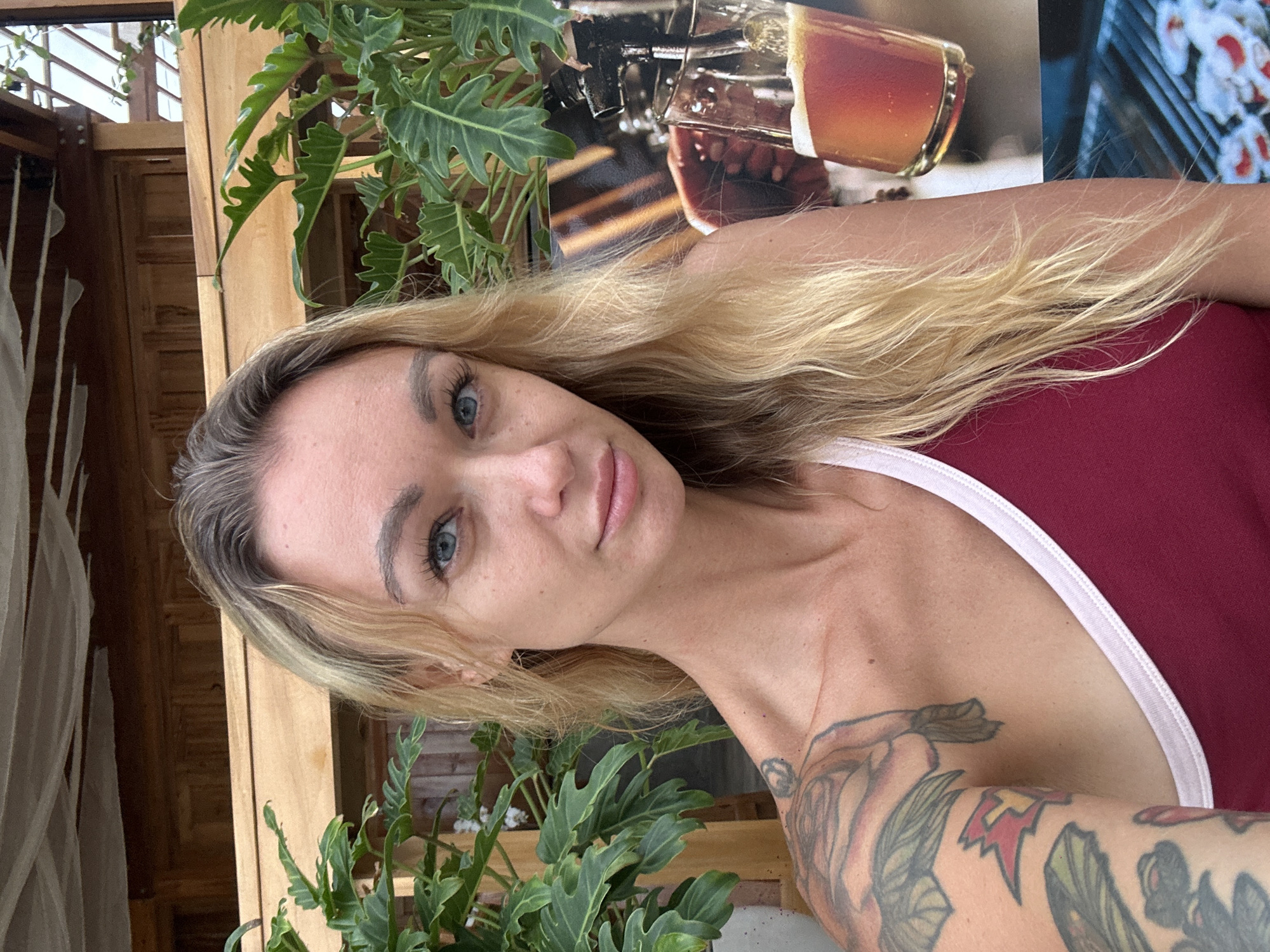 A selfie is taken by a woman with blonde hair and blue eyes, wearing a maroon and white tank top. She has tattoos on her left arm and shoulder. She is outdoors with a wooden structure and green plants behind her. To her right, there is a framed image of a glass of beer and a grill with food on it. The scene appears to be located in Phu Thuy Ward, Vietnam.