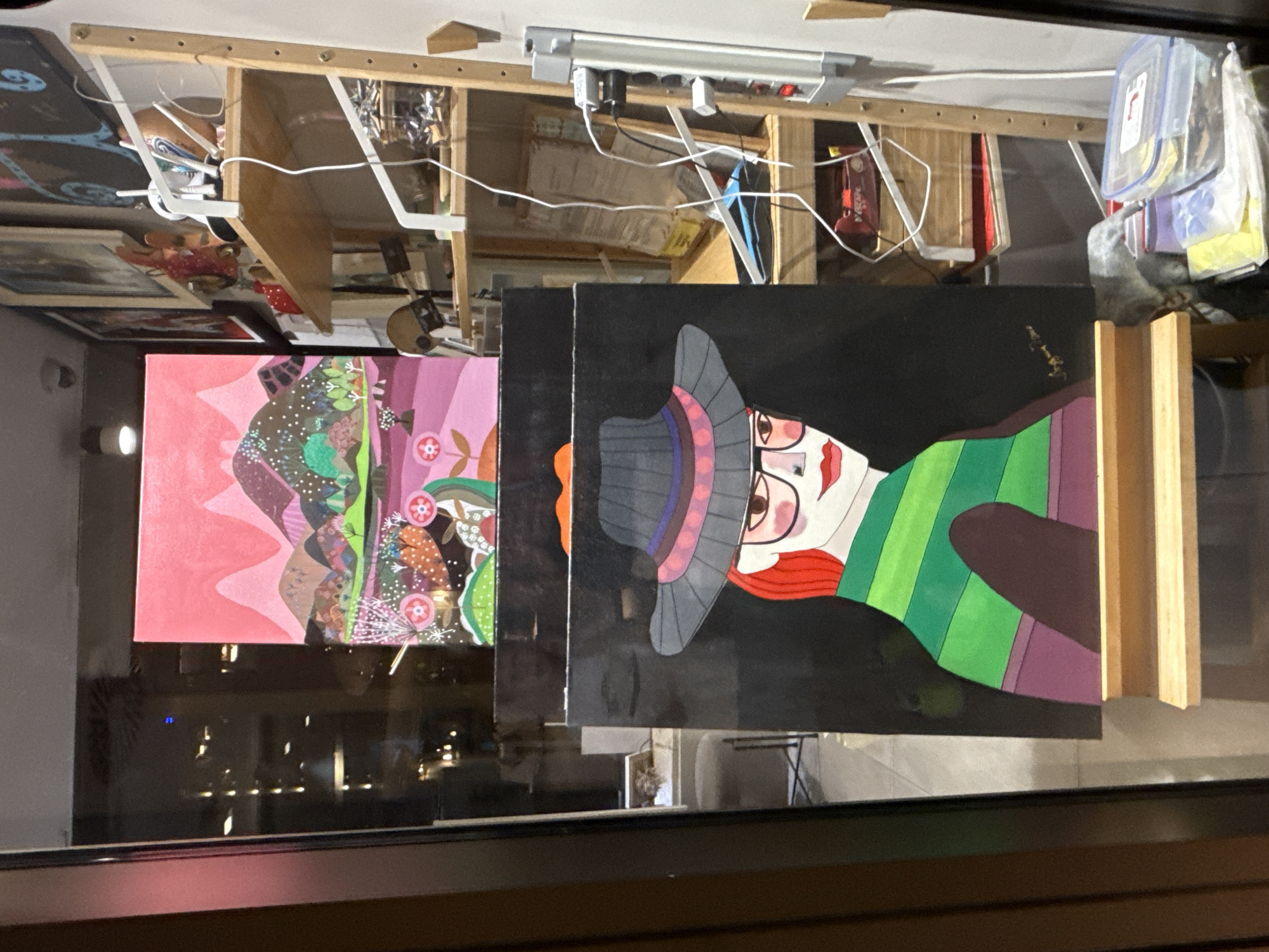 The image shows a display of artwork in a window. In the foreground, there is a painting of a woman with red hair, glasses, and a wide-brimmed hat. She is wearing a green and purple striped shirt. Behind this painting, there is another painting of a colorful landscape with pink mountains. The artwork is displayed on wooden easels, and shelves with various items are visible behind the paintings. The overall scene appears to be in an art shop or gallery. The specific location in Budva, Montenegro, is not discernible from the provided image.