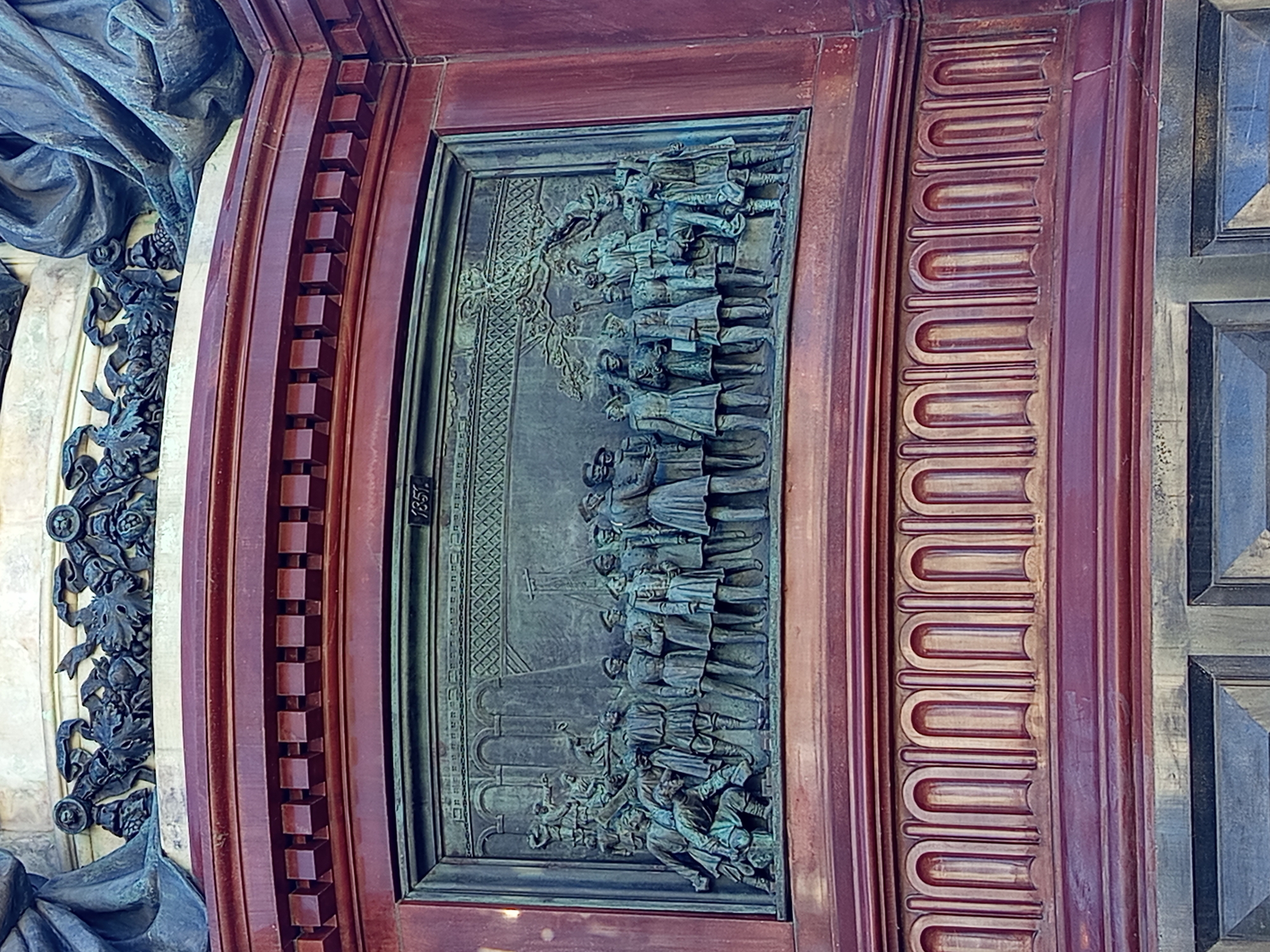 The image shows a close-up of a bronze bas-relief on the base of a monument in Saint Petersburg, Russia. The bas-relief depicts a group of people in historical attire, appearing to be engaged in some sort of assembly or ceremony. Some individuals are standing, while others are seated or kneeling. The scene is set against a backdrop of architectural elements and a stylized tree. In the upper right corner of the bas-relief, the year "1851" is inscribed. The bas-relief is framed by a dark, ornate border and is mounted on a large, dark red cylindrical structure, likely part of a monument's pedestal. The overall style suggests a commemorative or historical artwork.