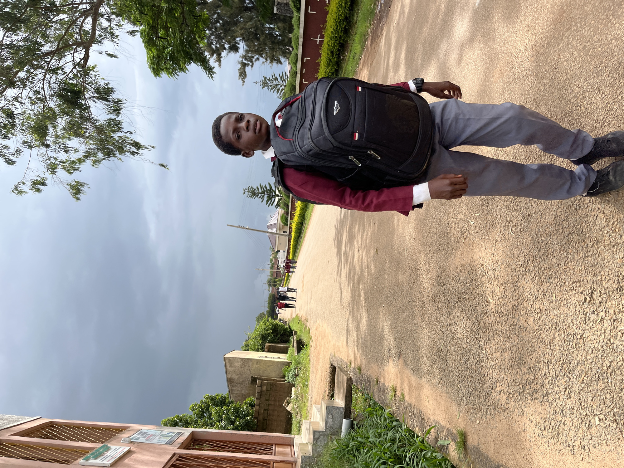A young boy stands facing the camera, wearing a maroon school uniform jacket over a white shirt, grey trousers, and dark shoes. He carries a large black backpack. The boy is positioned on an asphalt road in what appears to be a school compound or residential area, indicated by the presence of buildings, a fence, and trees. In the background, other students are walking away from the camera down the road, suggesting they might be going to or from class. The sky is overcast with dark clouds, hinting at an impending storm or recent rain. The lighting suggests it is daytime, possibly late afternoon. The setting is outdoors, with green foliage visible on either side of the road. There is no discernible text in the image. The boy's expression is neutral. The overall scene depicts a common day at school, perhaps during a break or after classes.