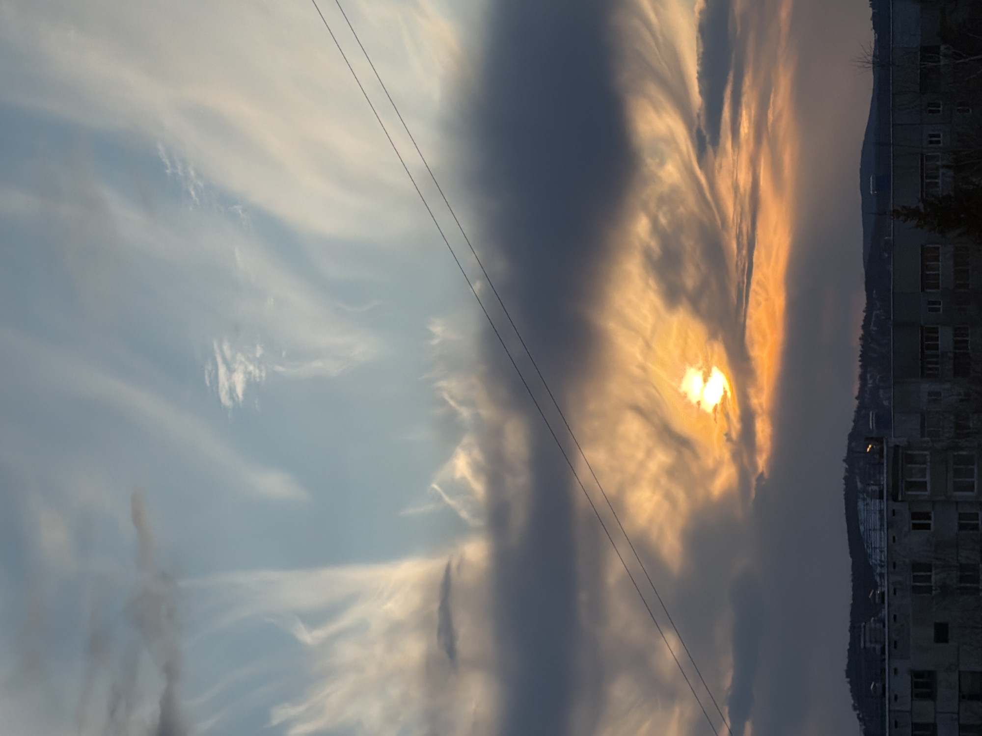 The media file captures a dramatic sunrise or sunset over a landscape in Ust-Ilimsk, Russia. The sky dominates the frame, presenting a dynamic interplay of light and clouds. On the left side, the sky is a pale blue, adorned with delicate, wispy white clouds, indicative of cirrus formations. Towards the center and right, the sky transitions to darker, denser clouds, which are intensely illuminated by the low-lying sun.
The sun, a brilliant white-yellow orb, is partially obscured by these clouds, casting a strong golden-orange glow that radiates through the cloud formations, creating an atmospheric and almost ethereal effect. Two dark, thin power lines cut diagonally across the sky from the upper left to the lower right, traversing both the bright and shadowed cloud areas.
Along the horizon, occupying the lower portion of the image, stands a dark, multi-story building with visible windows, suggesting a residential or industrial structure. Behind the building, a dark, low-lying hill or mountain range is visible, partially covered with what appears to be snow or frost, and sparse, bare trees dot the landscape, particularly near the building and on the hill. The overall scene suggests a cold climate.
The time of day is clearly either sunrise or sunset, known as the golden hour, due to the low angle of the sun and the warm, intense light it casts. The weather is partly cloudy, with a mix of high, wispy clouds and denser, mid-level formations. The urban-like building combined with the natural hilly landscape and bare trees provides a clear context for a town like Ust-Ilimsk. There is no visible text in the image.