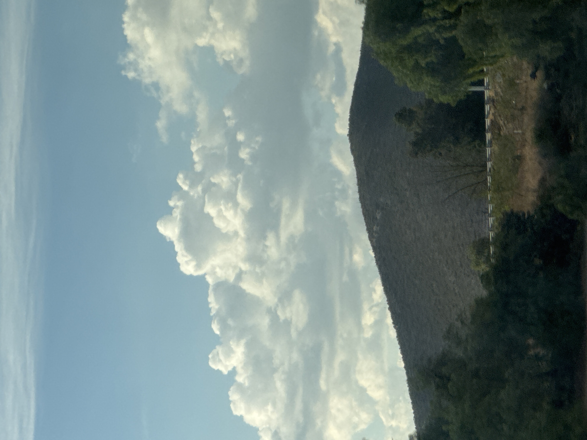 The image shows a landscape in Cuacnopalan, Mexico, featuring a combination of natural and man-made elements under a partially cloudy sky. The left side of the frame is dominated by a bright blue sky with wispy clouds. To the right, a large, dark gray hill or mountain extends vertically. A road and a white guardrail can be seen along the hillside, flanked by green trees and vegetation. The scene is observed during daylight.