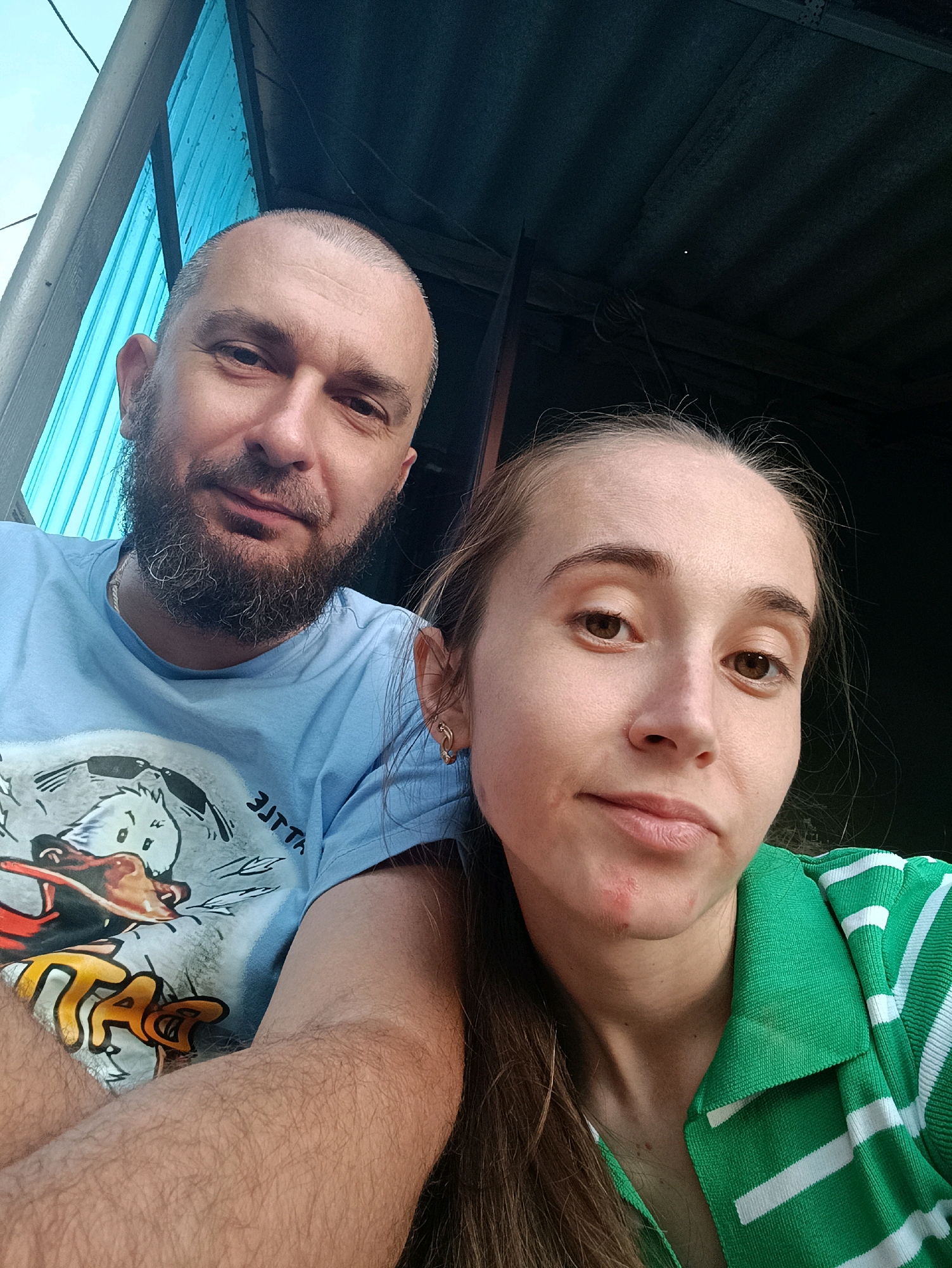 A man and a woman are posing for a selfie outdoors, with a corrugated metal roof visible above them and a blue corrugated plastic wall to their left. The man, who is balding with a beard, wears a light blue t-shirt with a cartoon print. The woman, with long brown hair, is wearing a green and white striped shirt. Both individuals are looking at the camera. The scene appears to be in Vypasne, Ukraine, though no specific landmarks are identifiable within the frame.