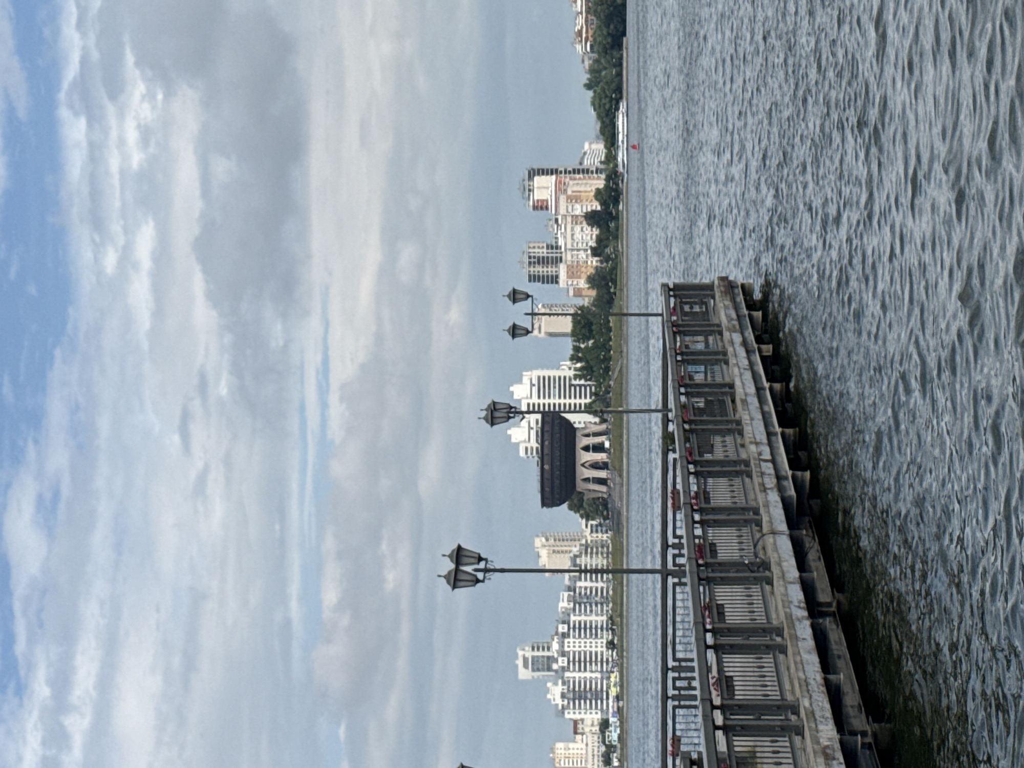This is a medium shot, taken from a low angle, of a pier extending into a body of water, with a cityscape in the background. The pier, made of weathered wood and metal railings, is lined with lampposts. The water is choppy and dark, with ripples visible. In the background, a modern city skyline features a cluster of white, tall buildings and a distinctive, large, dark bowl-shaped structure with arched openings at its base. Trees and greenery dot the shoreline between the water and the buildings. A white boat is visible on the water in the distance. The sky is overcast with a mix of grey and white clouds, with patches of blue sky visible. The overall impression is one of an urban waterfront scene on a cloudy day. The distinctive bowl-shaped structure is the monument known as the "Symbol of Kazan" or "Kazan Family Center", located in the city of Kazan, Russia.