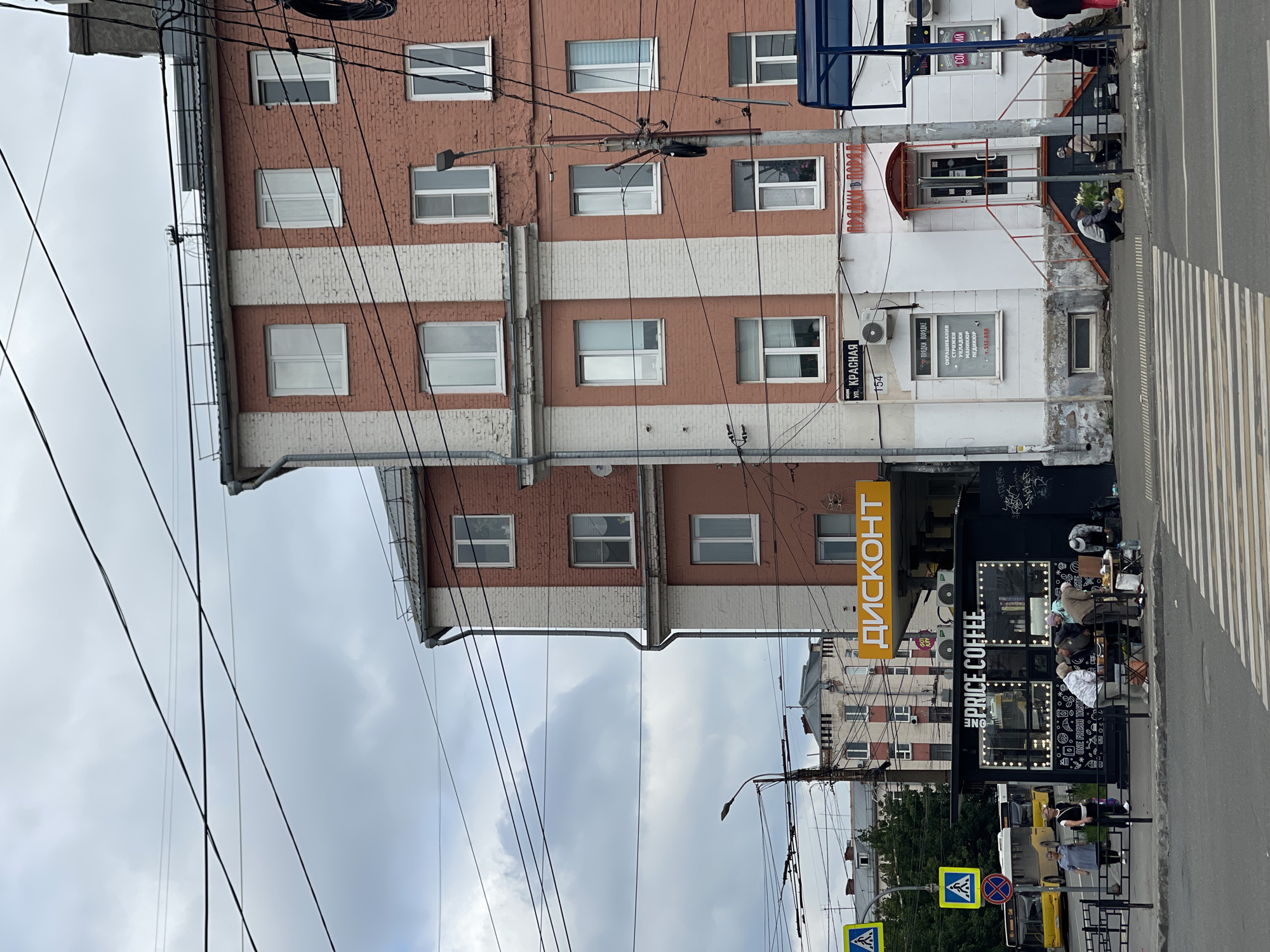 The image shows a street scene in Izhevsk, Russia, on a cloudy day. In the foreground, there is a crosswalk. To the left, a black kiosk with "PRICE COFFEE" illuminated in lights and an advertisement for "ONE FRESH TOAST" is set up. Several people are gathered around the kiosk, some interacting with each other and others seemingly making purchases or examining items on a table.
Behind the kiosk, a multi-story building with red brick and white accents is visible. A bright orange sign with the word "ДИСКОНТ" (Discount) is prominently displayed on the building above the kiosk. Further up the street, another building with a similar architectural style can be seen. Power lines crisscross the sky, adding to the urban environment.
To the left of the kiosk, a yellow bus is parked near a pedestrian crossing sign. More people are visible walking on the sidewalk and near the bus. The building on the right has several windows and air conditioning units. A blue awning extends from the right side of the image. The overall impression is a bustling urban street with commercial activity and pedestrian traffic.
