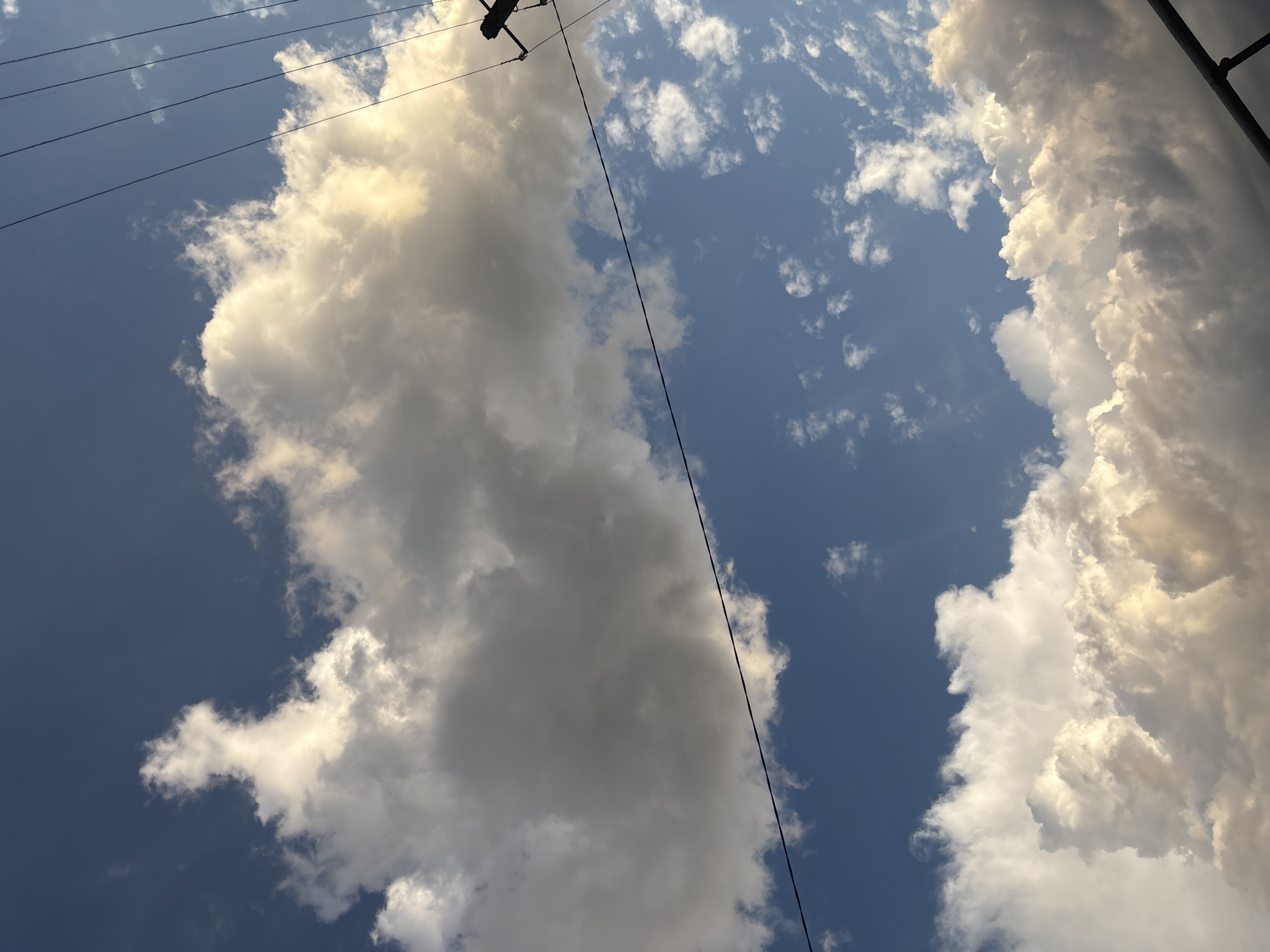 This media file depicts a dramatic sky filled with large, billowy clouds. The clouds are illuminated by sunlight, with some areas appearing bright white and others in shadow, creating a sense of depth and texture. The sky itself is a deep blue, indicative of a clear day with scattered clouds.
Thin, dark lines, which appear to be power lines, stretch diagonally across the frame. These lines, along with a hint of a pole or structure at the top edge, suggest an outdoor setting with man-made infrastructure. The angle of the shot, looking upwards, emphasizes the vastness of the sky and the towering clouds.
The lighting suggests it is either early morning or late afternoon, as the sunlight is casting a warm glow on the clouds. The weather appears to be fair, with fluffy cumulus clouds that are not indicative of imminent storms.
While the specific location is Ladozhskaya, Russia, there are no overt visual cues within the image to confirm this beyond the provided context. The overall impression is one of natural beauty and the grandeur of the sky. There is no visible text in the image.
