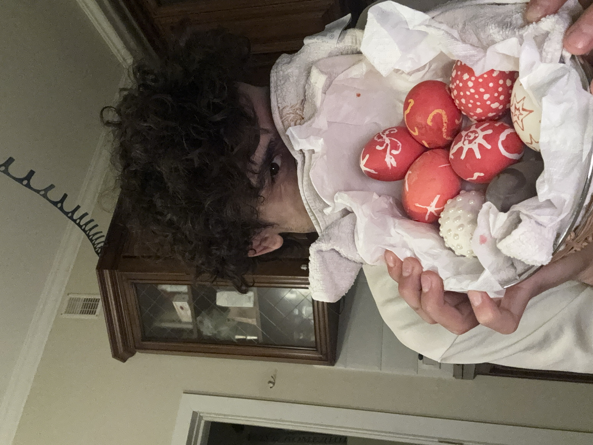 A person with curly dark hair holds a metal bowl filled with decorated Easter eggs and crumpled white paper. The eggs are primarily red with white designs; one eggshell is patterned with red polka dots on a white background. The bowl also contains a decorative pearl-like sphere and what appears to be a gray stone.
The person is indoors, standing in front of a dark wood cabinet with a glass door. The wall is light beige. There is a white door frame and part of a white door visible near the bottom of the image. A black clothes hanger is mounted on the wall in the upper left of the frame.
There is no information in the image to support a location in Moscow, Russia.