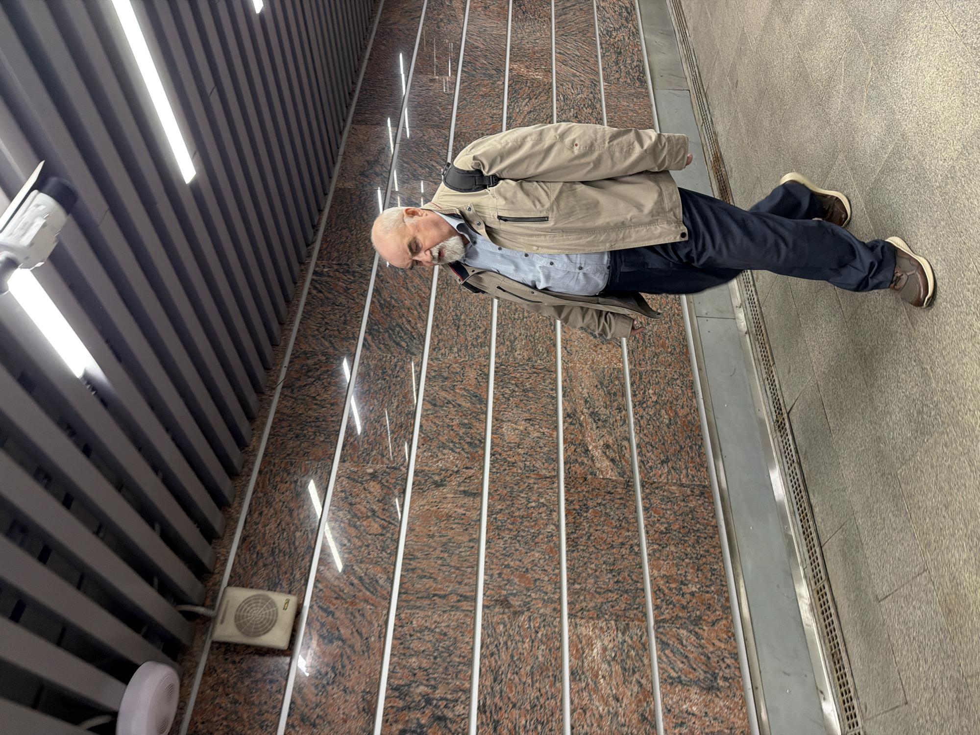 An elderly Caucasian man with a gray beard is walking through a modern underpass or subway station in Moscow, Russia. He is wearing a light brown jacket over a blue collared shirt, dark blue trousers, and brown athletic shoes. A black backpack is visible on his shoulders. The underpass features a granite-patterned wall with horizontal white stripes and a metal grate along the bottom. The ceiling is constructed of gray metal slats with illuminated fluorescent lights. A white speaker and a white security camera are mounted on the ceiling. The floor is paved with gray stone tiles. The man is walking from left to right in the frame, looking down and to his left.