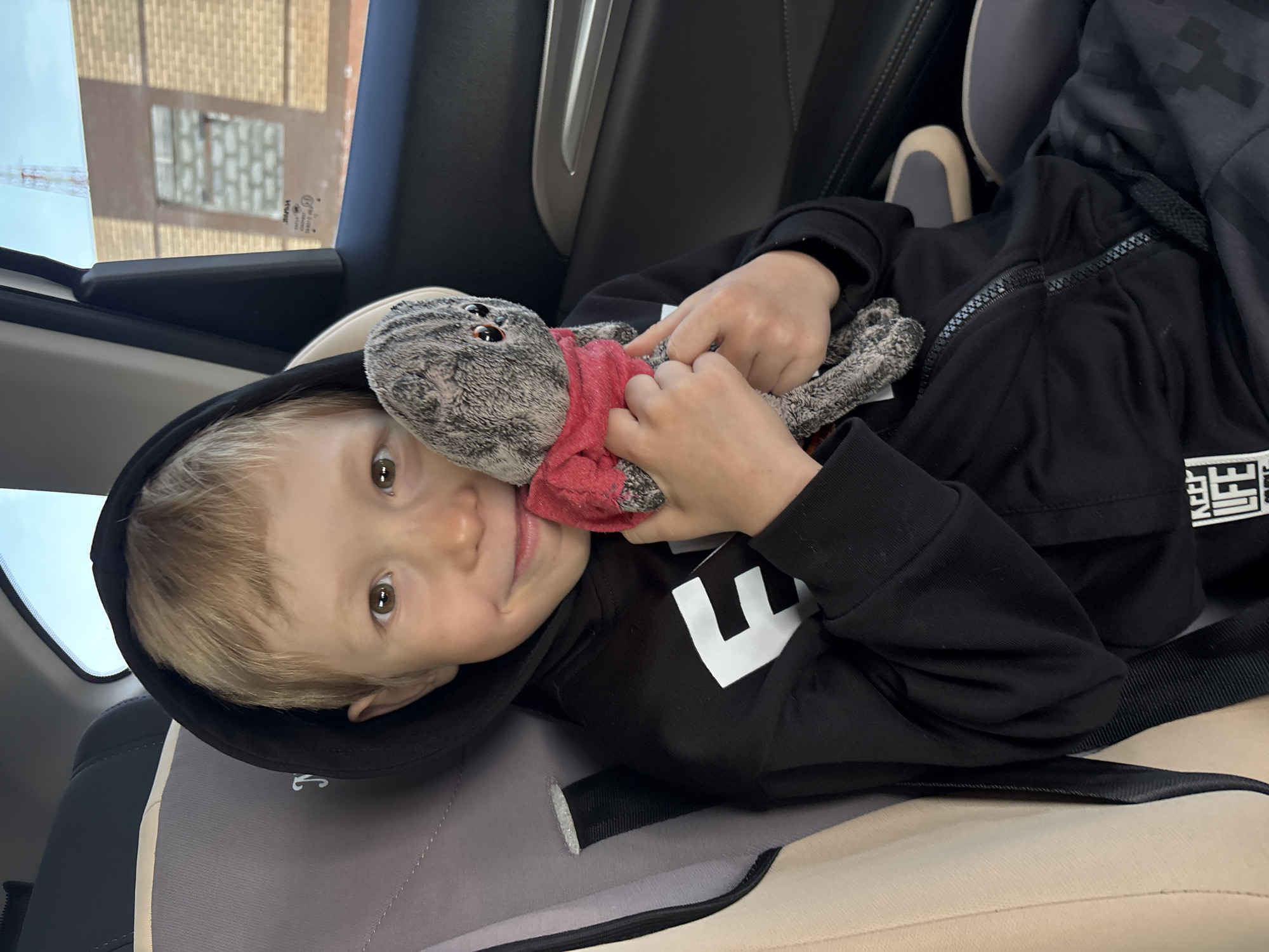 A young child is lying in a car seat, holding a stuffed cat toy. The child is wearing a black hooded sweatshirt with white lettering. The car is in Chaikovsky, Russia, as can be inferred from the writing visible through the window.