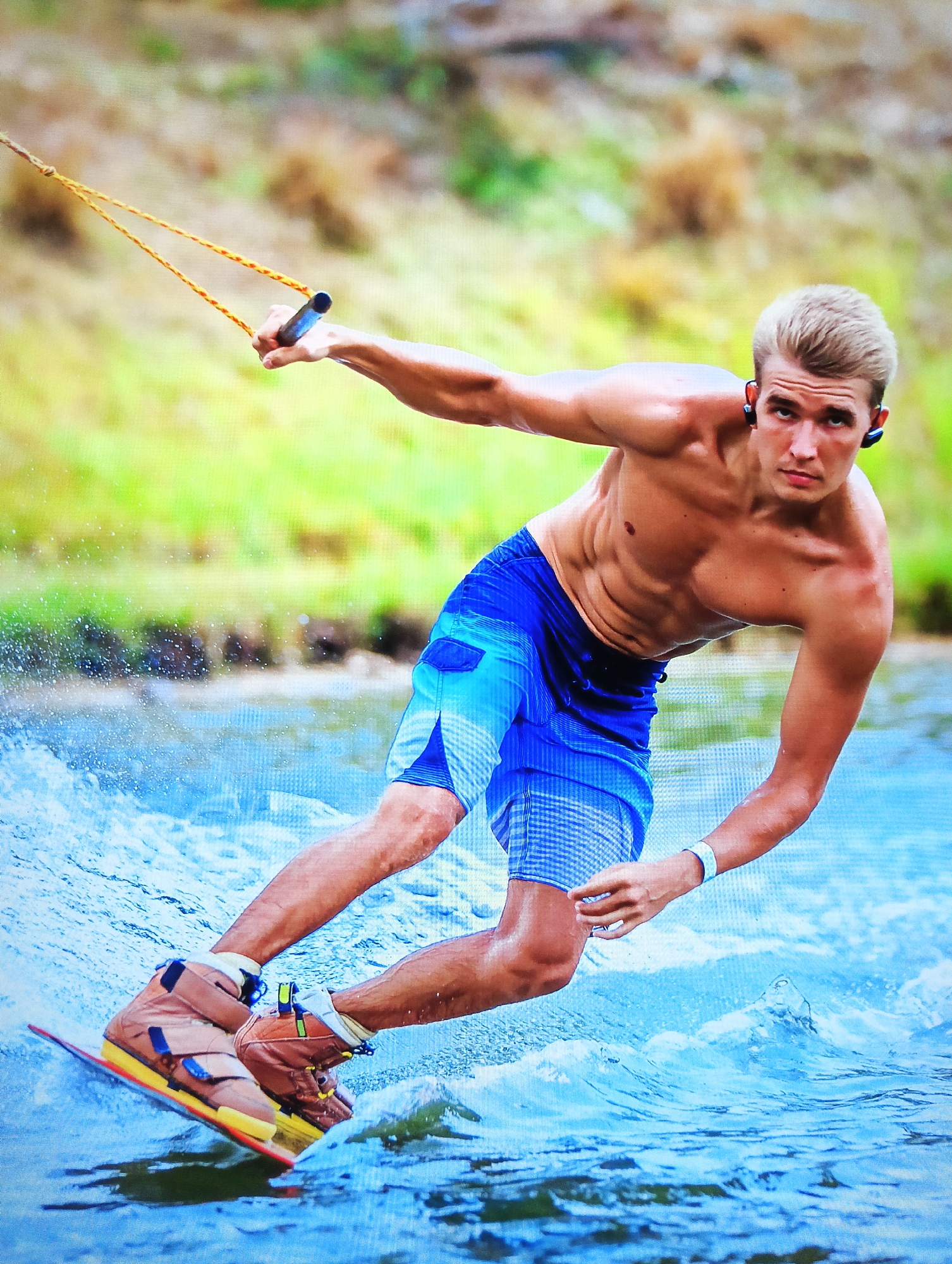 A man wakeboards on a body of water. The man is shirtless and wearing blue shorts, wakeboard boots, and wireless earbuds. He is holding a wakeboarding rope with his right hand. The man has light skin, blonde hair, and is looking at the camera. Water splashes around the wakeboard. In the background, green vegetation can be seen. The man is located in Nha Trang Ward, Vietnam.