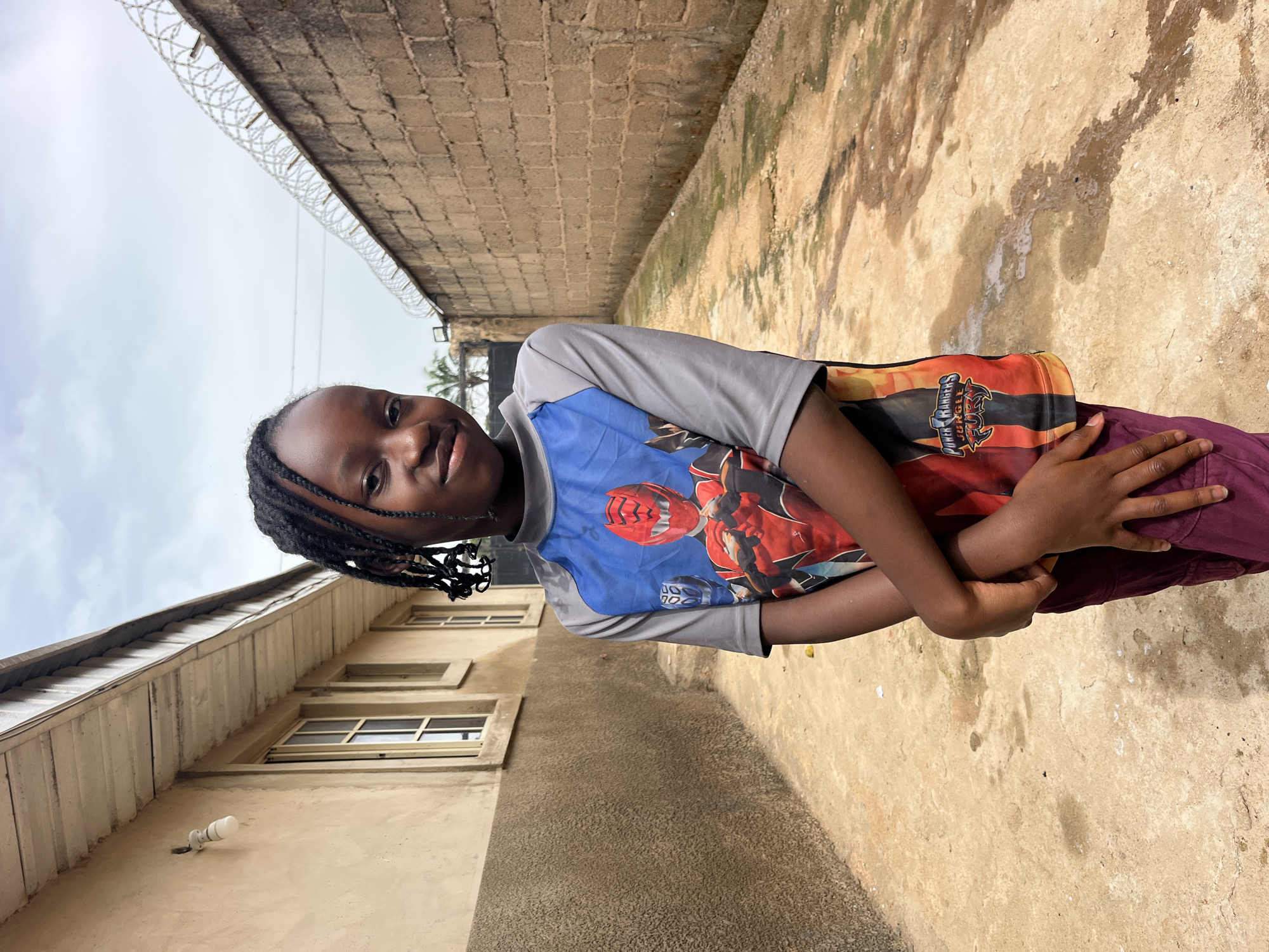 A young Black girl with braided hair stands in an alleyway between two buildings. She is wearing a gray t-shirt with a Power Rangers graphic on the front and maroon pants. The girl is looking at the camera with a slight smile. The alleyway is paved with concrete and has a wall on the right side with barbed wire on top. The building on the left is a light brown color with windows. The sky is cloudy.