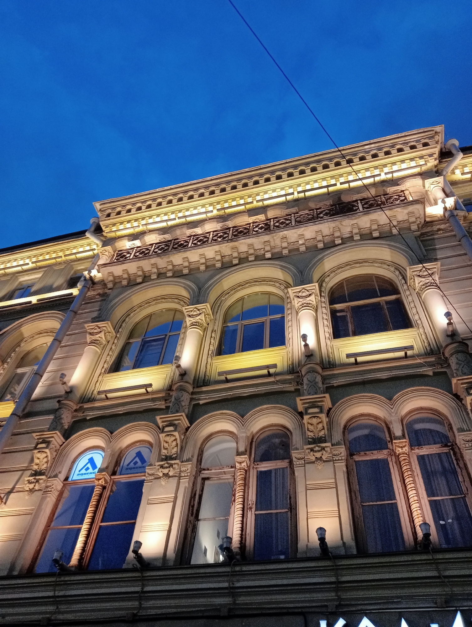 An upward angle shot of an ornate building facade illuminated by yellow lights against a dark blue twilight sky. The building features multiple levels of arched windows, decorative columns, and intricate architectural details, including a balcony with a patterned railing. A drainpipe runs vertically down the left side of the facade. Two windows on the second floor from the bottom display a blue illuminated logo with a stylized "A" and Cyrillic text. The sky is clear with a faint, diagonal power line stretching across it. The image appears to be taken in Saint Petersburg, Russia, given the architectural style common in the city.