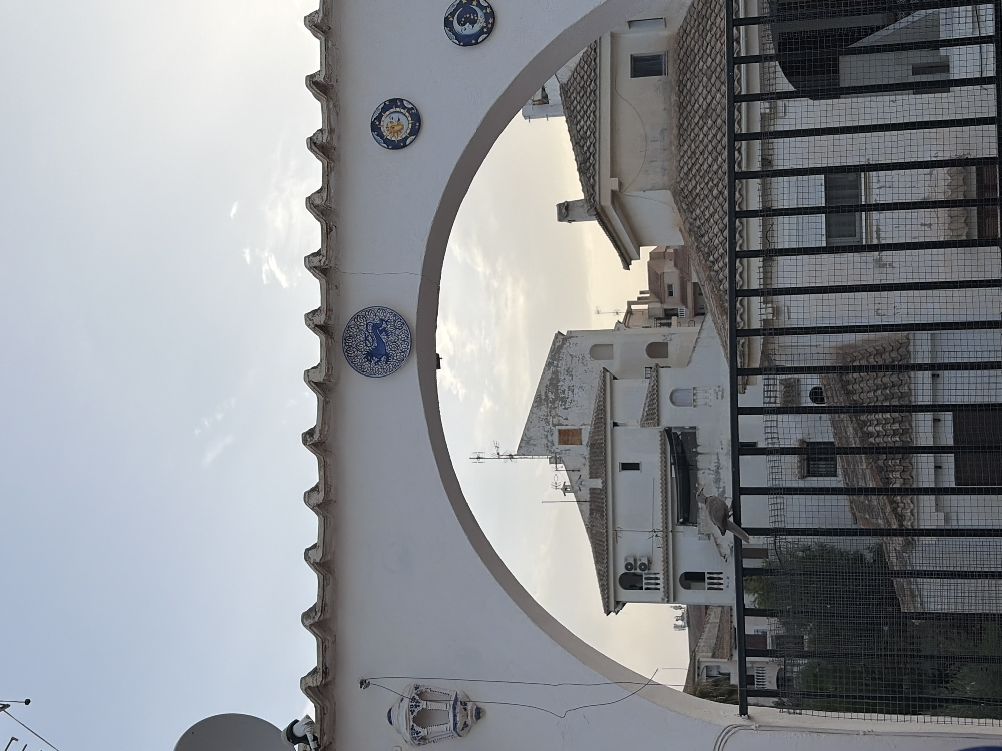 An arched white structure with a tiled roof spans the foreground, adorned with a decorative lantern on the left and three circular ceramic tiles featuring celestial bodies and a mythical creature on the right. Through the archway, a bird is perched on a black metal fence. Beyond the fence, white buildings with tiled roofs and balconies are visible under a pale sky. The scene suggests a view from a rooftop or elevated position in a Spanish town, likely Torrevieja, characterized by its whitewashed architecture and Mediterranean climate.