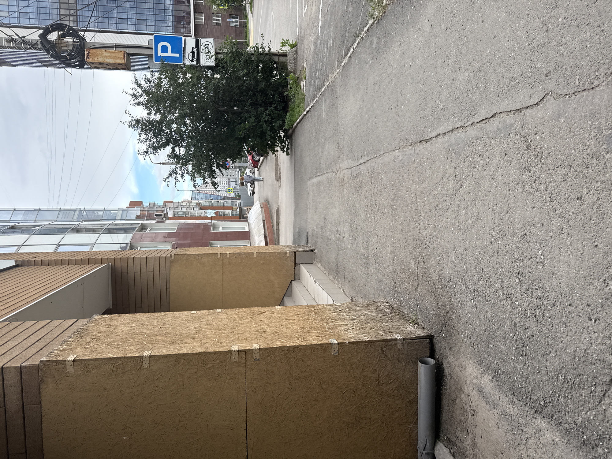 The image depicts a sidewalk and street scene, likely in Perm, Russia, given the context. A large, unfinished building with wood paneling covers the left side of the frame, obscuring the lower portion of a brick and glass structure. The sidewalk is made of grey asphalt with visible cracks and is slightly uneven. To the right, a tree grows adjacent to the sidewalk, and a metal pole stands nearby. On this pole, a blue parking sign is visible, accompanied by a sign with a car icon and numbers "10 15 20". Further down the street, several cars are parked, and other buildings, including a modern glass-fronted one, can be seen. The sky above is overcast with patches of blue. A grey pipe lies horizontally on the ground near the base of the wood-paneled building. A person, dressed in light-colored clothing, is walking away from the camera on the sidewalk in the distance. The scene suggests an urban environment, possibly undergoing some form of construction or renovation.