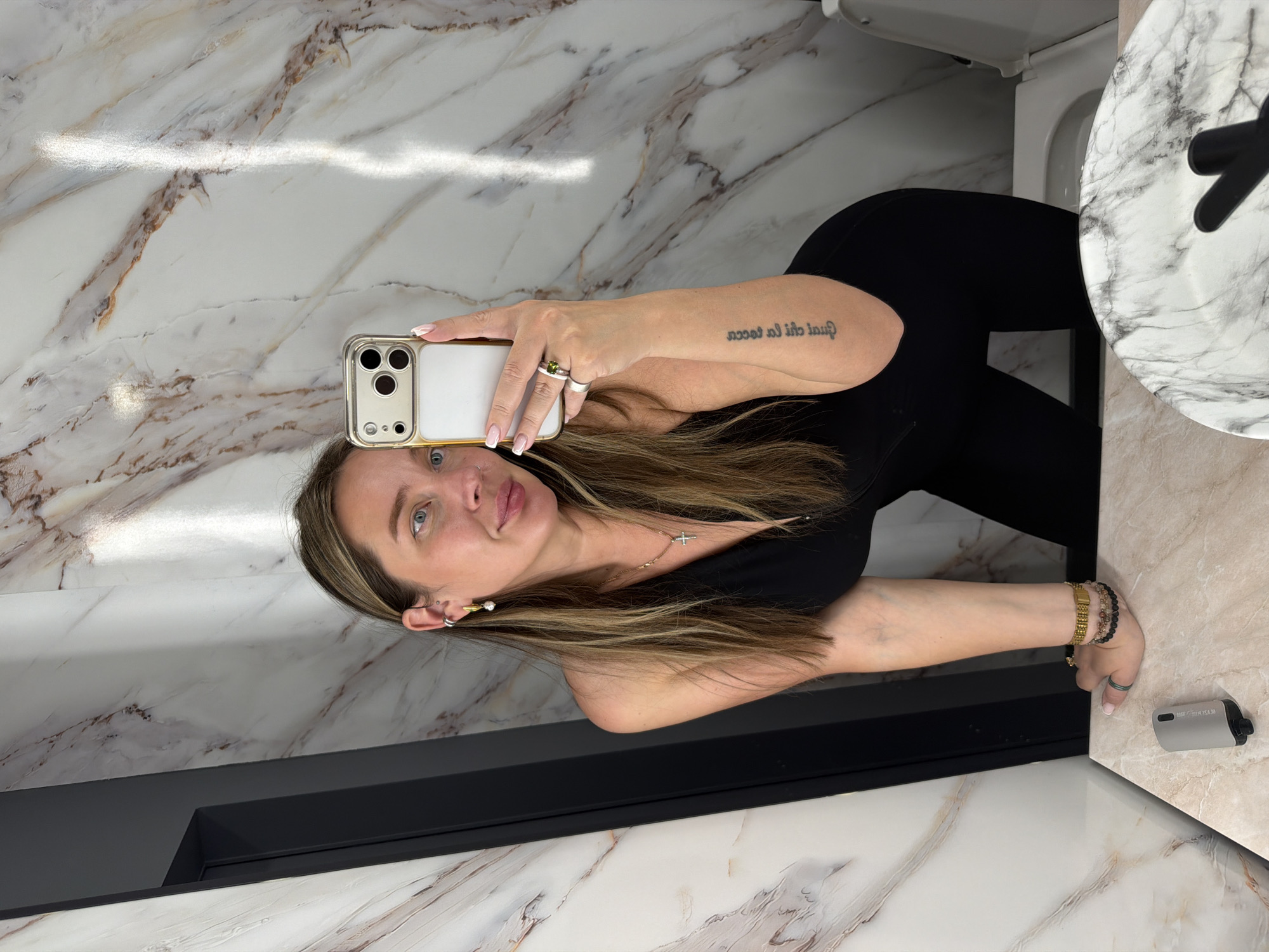 A woman is taking a selfie in a bathroom. She is wearing a black top and leggings. She is holding a smartphone with a gold case. The bathroom has marble walls and a marble countertop. There is a toilet and a sink. The location appears to be a bathroom, and there is no information suggesting this is located in Moscow, Russia.