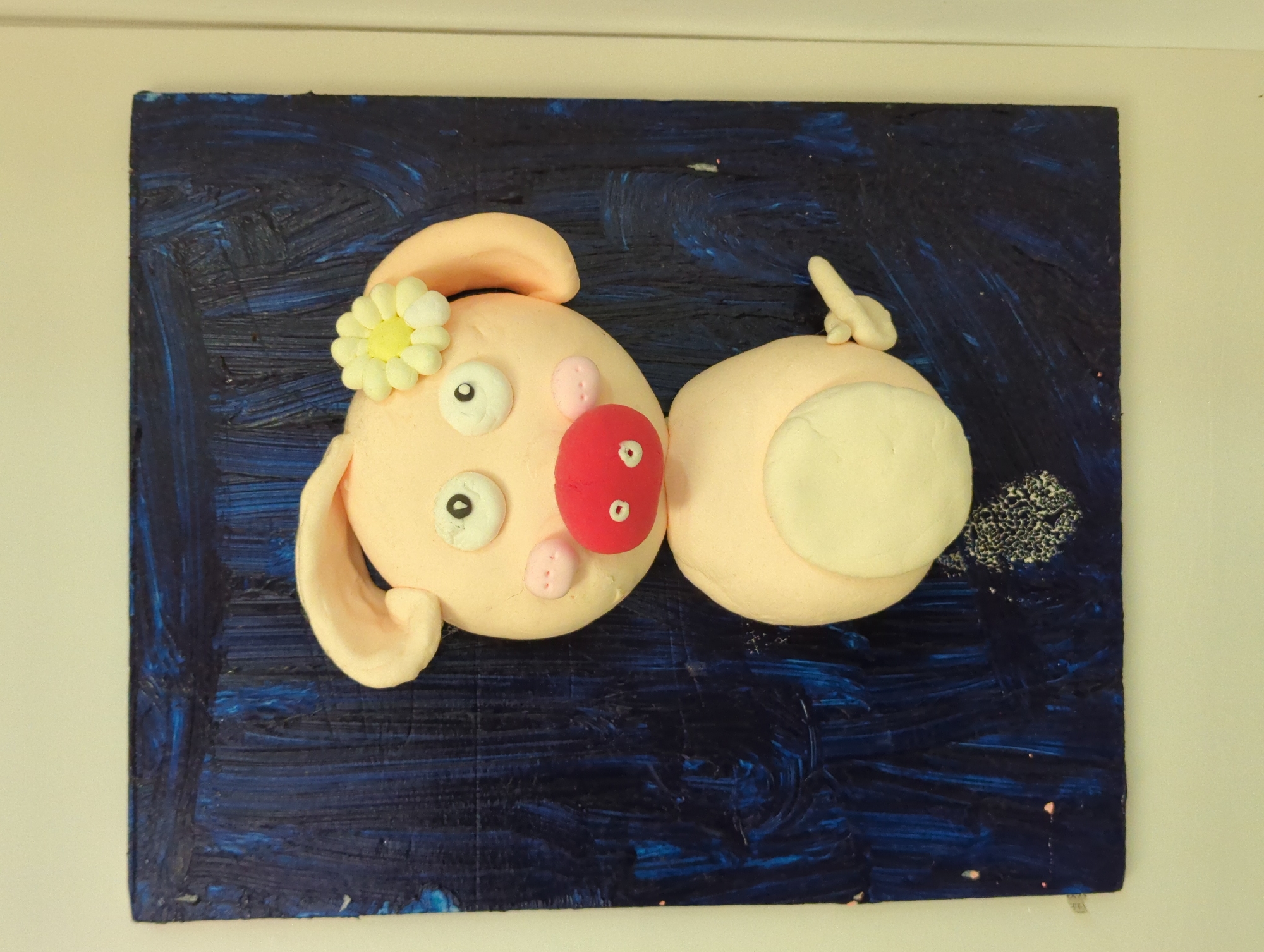 The image shows a clay pig sculpture on a dark blue, textured, square background. The sculpture is a light pink color with a round head and body. The pig has a red nose, white eyes with black pupils, two pink dots for nostrils, and a white flower behind its ear. The pig's tail is curled up and is white with a pink tip. There is a dark, textured spot near the pig's tail. The image appears to be taken indoors with a light wall visible behind the sculpture. There is no evidence in the image that indicates that this image was taken in the city of Shunde District, China.