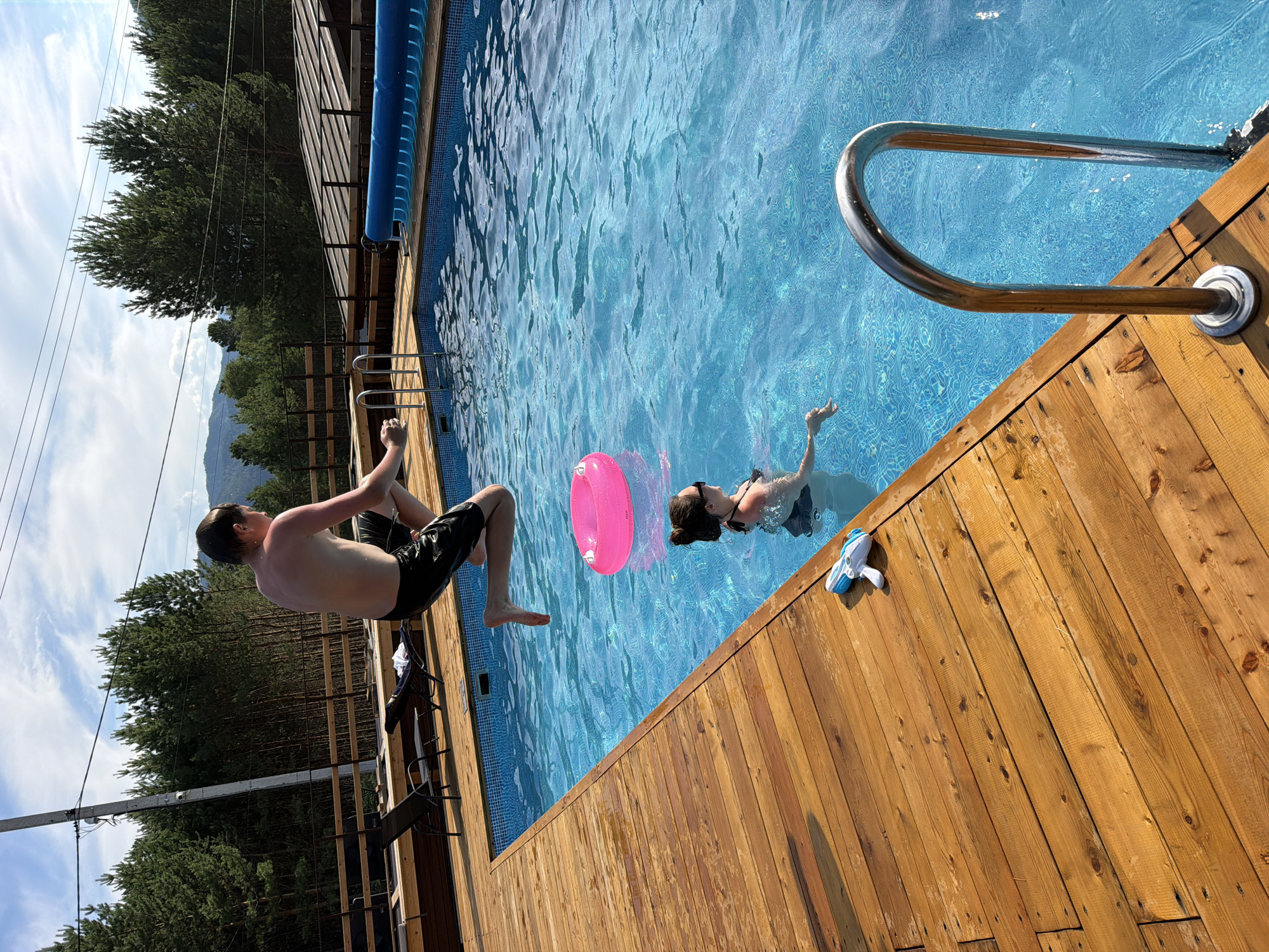 In Elekmonar, Russia, a young male in black shorts is captured mid-air as he jumps into a bright blue swimming pool. He is positioned above and to the left of a woman who is swimming in the pool. To the woman's right, a pink inflatable ring floats on the surface of the water. Nearby, on the wooden deck of the pool, a white and blue water gun rests on the planks. The scene is set outdoors, with a lush forest of tall pine trees bordering the pool area. The sky above is partly cloudy, with a hint of mountains visible in the distance. A wooden fence and structures are also present around the pool.