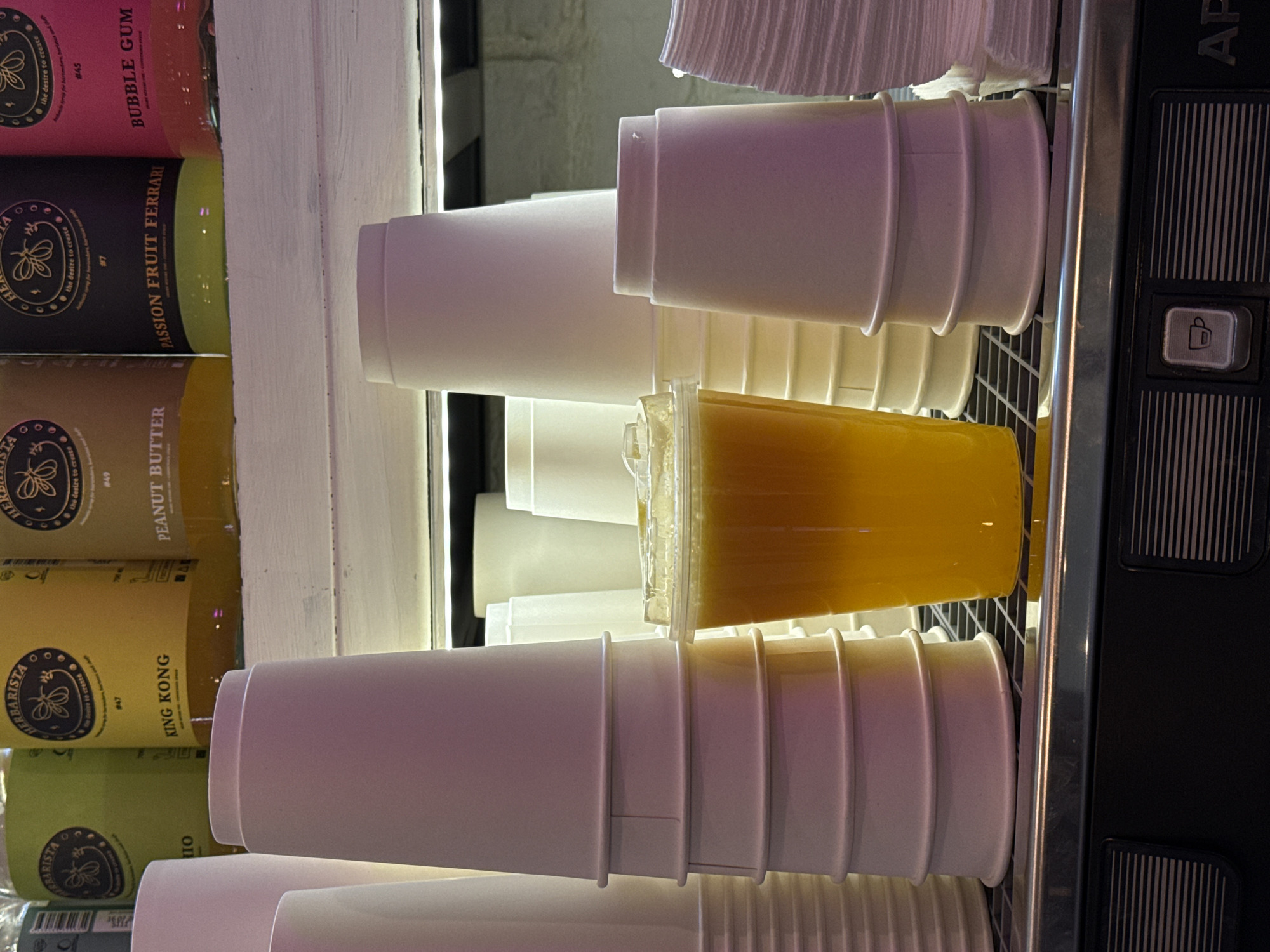 This image captures a close-up view of a beverage display, likely in a cafe or bar. In the foreground, a clear plastic cup filled with a bright orange beverage is prominently featured. The cup is resting on a dark metal grate. To the left of the beverage, a tall stack of white paper cups is visible, with distinct ridged rings around their exteriors. To the right of the beverage, another stack of white paper cups, slightly shorter and with similar ridging, is present. Behind these cups, in the mid-ground, are additional stacks of white paper cups of varying heights. In the background, several tall bottles with colorful labels are arranged on a shelf. These bottles have circular logos with insect imagery and text that appears to be in English. Specifically, the labels read "HERBARISTA," "KING KONG," "PEANUT BUTTER," and "PASSION FRUIT FERRARI," and "BUBBLE GUM". The lighting in the scene appears to be artificial, with a slight purple and pink hue cast on the cups, possibly from ambient lighting or a nearby light source. The overall environment suggests a commercial establishment preparing for or serving drinks. The image does not provide any specific details or context to pinpoint its location within Moscow, Russia.