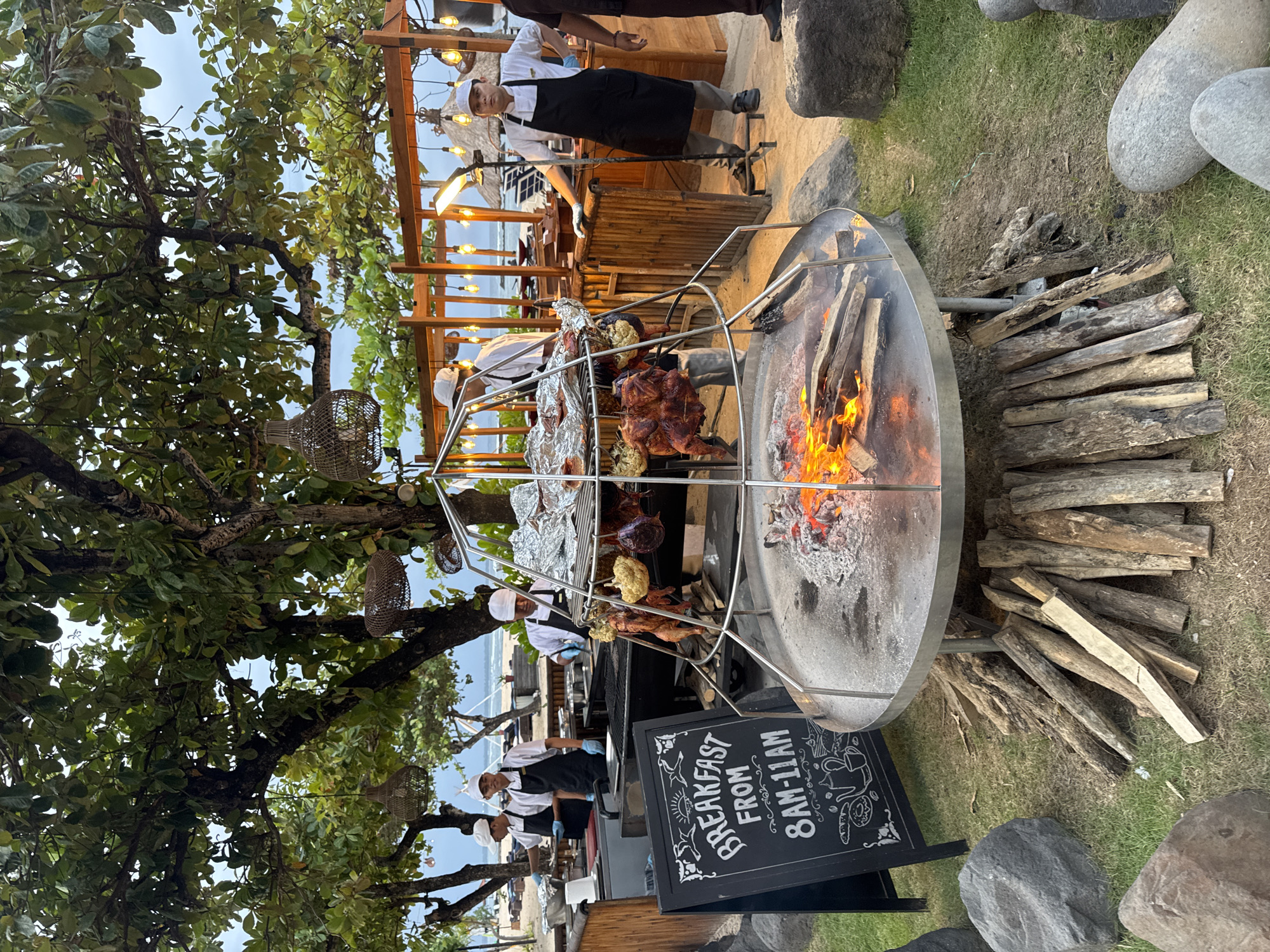 The image captures an outdoor cooking scene at what appears to be a restaurant or resort, possibly in Kuta Selatan, Indonesia. A large, circular, open-flame grill is the focal point, with a metal structure holding foil-wrapped items and roasted chickens suspended over the fire. A chef in a white uniform and black apron is standing nearby, seemingly attending to the cooking process.
Next to the grill is a chalkboard sign announcing "Breakfast from 8 AM - 11 AM" with decorative drawings. Piles of firewood are stacked around the base of the grill.
The setting includes a partial view of an outdoor dining area with wooden structures and string lights. In the background, there is a glimpse of a beach and ocean. A large tree with green foliage provides shade and adds to the outdoor ambiance. Several people in chef uniforms are near the grill. The surrounding ground is covered with grass and large stones or boulders.