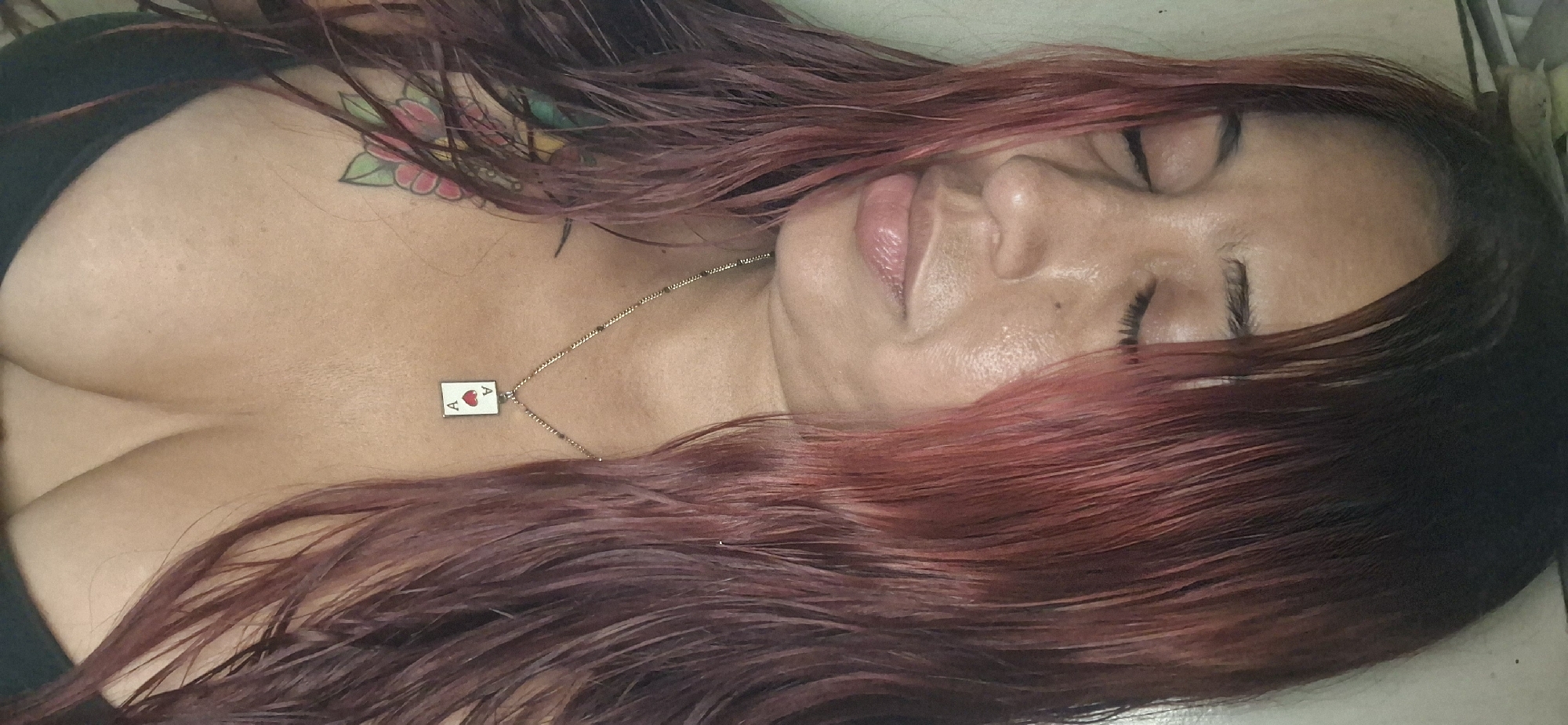 A woman with reddish-brown hair lies on a flat surface with her eyes closed. She is wearing a black top and a necklace with an "Ace of Hearts" pendant. A floral tattoo is visible on her upper chest/shoulder area. Her skin appears moist. There is no indication of the location being Vallecito, Venezuela in the image.