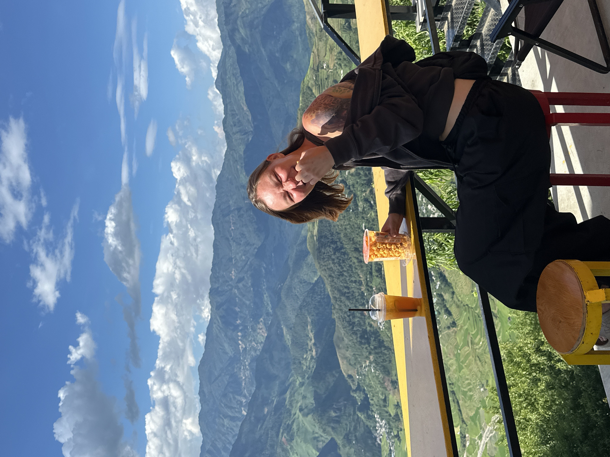 An adult female is seated at an outdoor establishment overlooking a vast mountainous landscape under a blue sky with scattered white clouds, consistent with the terrain found in Tu Le, Vietnam. She is positioned at a high yellow table or counter, facing slightly left towards the camera, and displaying a broad smile or laugh with her eyes squinted. Her right hand is raised to her mouth, holding a clear plastic cup filled with orange-colored popcorn. A clear plastic cup with a black straw, containing an amber beverage with fruit slices, rests on the table to her left. She wears a dark zip-up hoodie, partially pulled off her left shoulder, revealing extensive tattoos on her left shoulder and upper arm. She also wears a dark top, showing a sliver of her midriff, and dark trousers. She is seated on a red stool; another yellow-legged, wooden-topped stool is partially visible to the far left under the table. A black metal railing runs below the yellow table. The background features multiple layers of green, forested mountains with valleys, where scattered clusters of buildings and visible terraced fields indicate human settlement. The sunlight is bright, casting distinct shadows.