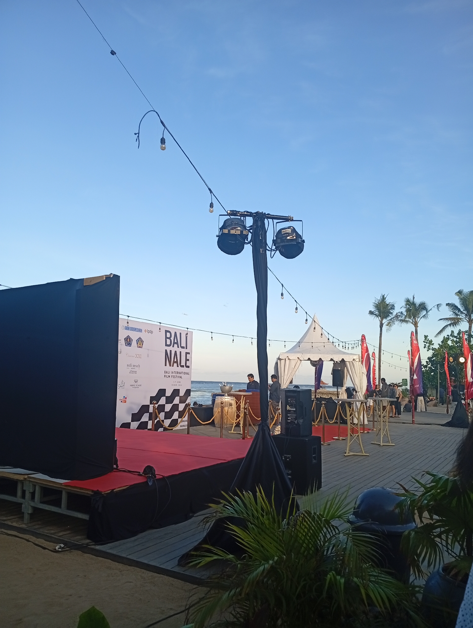 The image shows a stage setup at an outdoor event, likely a film festival based on the "BALI NALE BALI INTERNATIONAL FILM FESTIVAL" banner. The scene appears to be near a beach, with the ocean visible in the background. There's a red carpet leading to the stage, flanked by black curtains on one side and a banner with logos and festival branding on the other. A tent is visible in the background. Several people are standing or walking around the area, suggesting preparations or ongoing activity. Lighting equipment is prominent, indicating the event is likely planned for the evening. Palm trees are visible, suggesting a tropical location. The scene is in Denpasar, Indonesia.