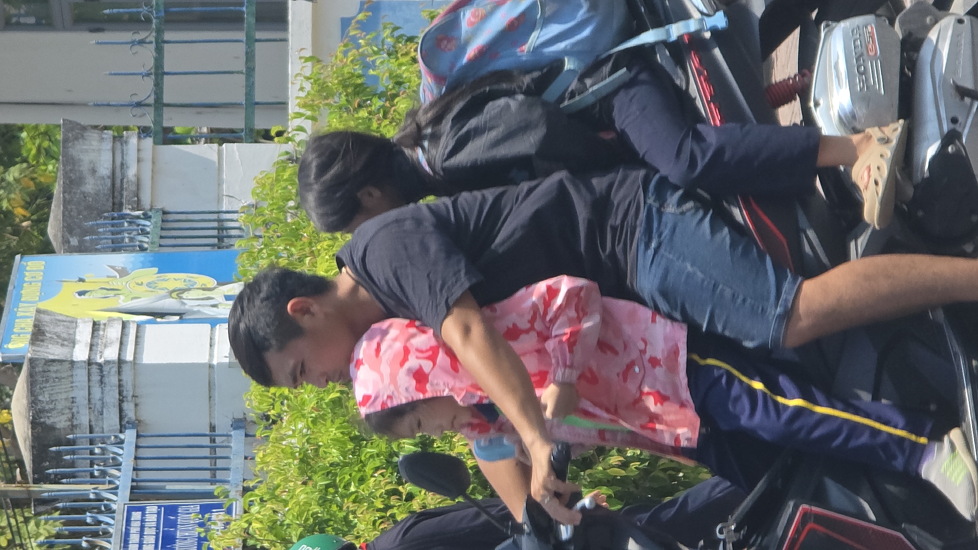 A family of three is precariously arranged on a parked motorcycle in Nha Trang, Vietnam. An adult male in a dark t-shirt and blue jean shorts is at the front, gripping the handlebars. A small child wearing a pink camouflage jacket and blue pants with a yellow stripe sits in front of him. Behind him is another child with long dark hair, wearing a dark jacket and pants with a blue backpack. They are positioned in front of a building with a sign that is partially visible. Vegetation is behind them.