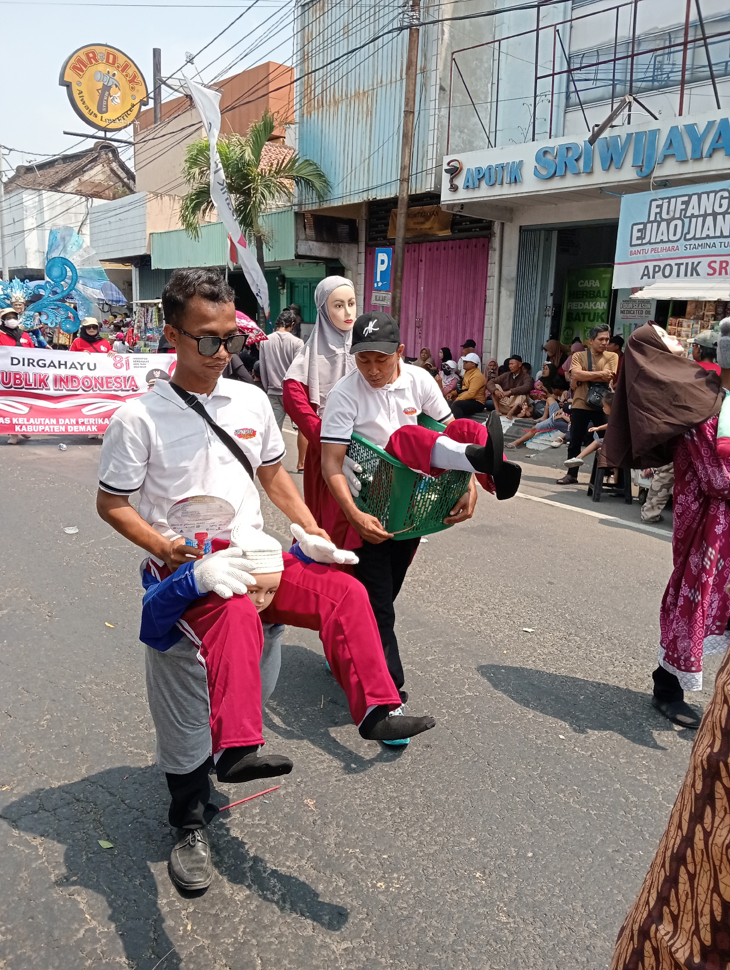 The image depicts a street scene in Demak, Indonesia, during what appears to be a parade or public event. In the foreground, two men are walking. The man on the left is carrying a mannequin torso, dressed in red pants and a blue and white top, with white gloves on its hands. The man on the right is carrying a green basket containing a mannequin dressed in red with white gloves. Both men are wearing casual clothing.

Behind them, other participants and spectators are visible. A banner with Indonesian text, including "DIRGAHAYU REPUBLIK INDONESIA", is stretched across the street. Several buildings line the street, one of which has a sign that reads "APOTIK SRIWIJAYA". People are seated on the roadside, watching the procession. The environment is urban, with paved roads and overhead electrical wires. The lighting suggests it is daytime, with clear skies and shadows indicating a sunny day.