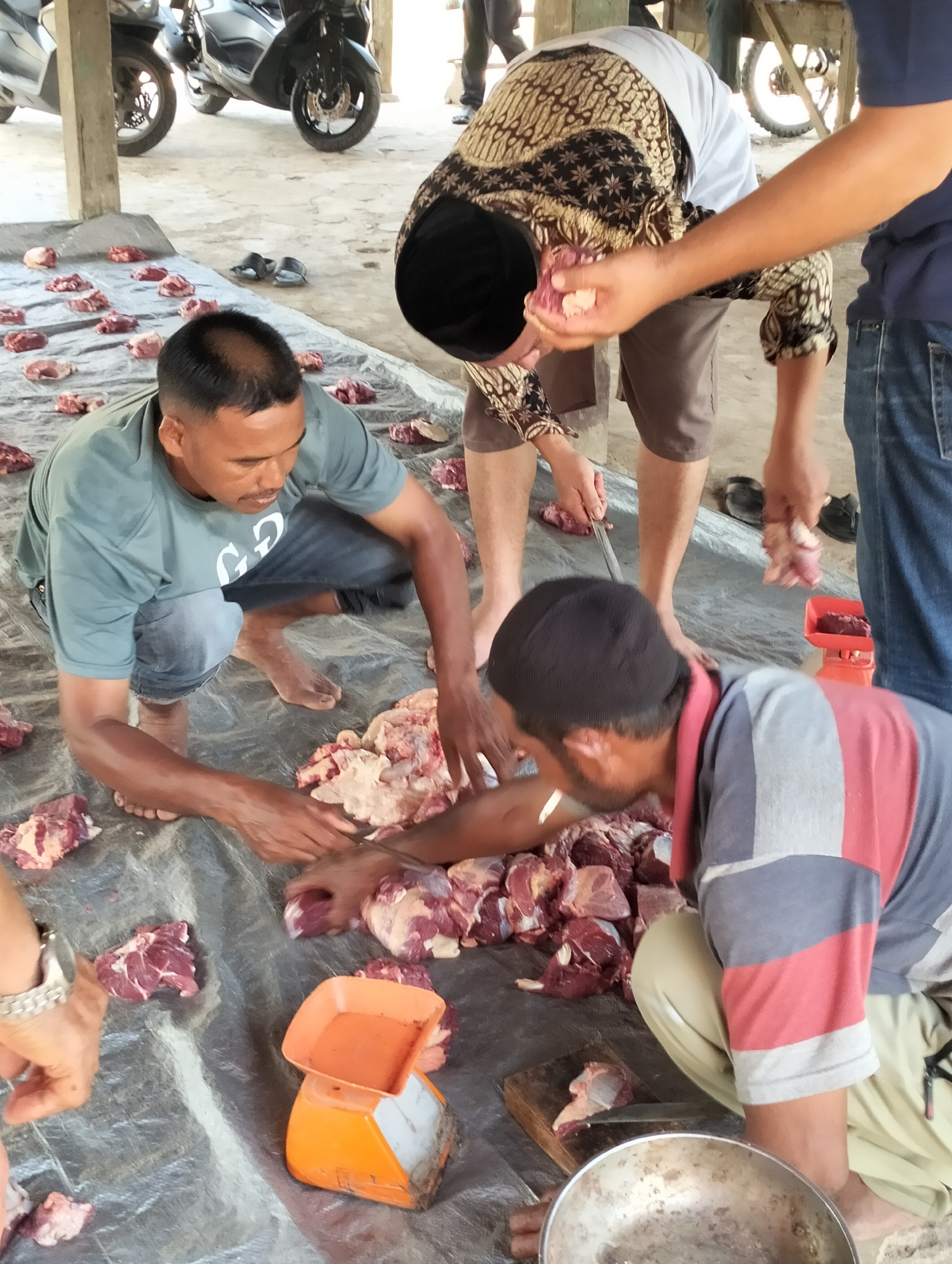 Men in Meunasahalue, Indonesia, are butchering meat on a large tarp under a covered structure. Several men are barefoot and squatting or kneeling around the tarp, which is littered with pieces of raw meat. One man in a green t-shirt is cutting meat with a knife. Another man wearing a patterned shirt is holding a piece of meat. An orange scale sits nearby. In the background are parked scooters and more people standing.