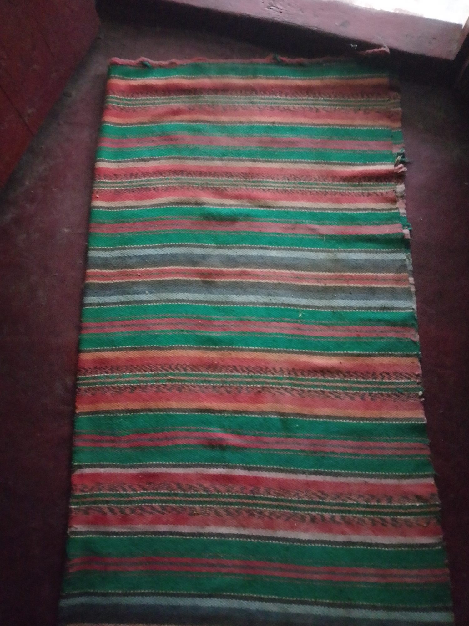 A rectangular, woven textile, approximately 1 meter in length, is laid flat on a dark reddish-brown surface, consistent with a floor, in the city of Unknown, Unknown. The textile features horizontal stripes of varying widths and colors, predominantly dark green and muted red/orange. Interspersed are thinner stripes of light grey, white, and dark blue/grey. The texture indicates a coarse weave, possibly wool or similar fiber. The top and right edges show minor fraying. Adjacent to the textile on the left is a darker, less defined reddish-brown surface, while the upper portion of the image reveals darker, blurred architectural elements, possibly a wall or baseboard. The overall lighting is dim and uniform. No individuals or active interactions are present in the scene.