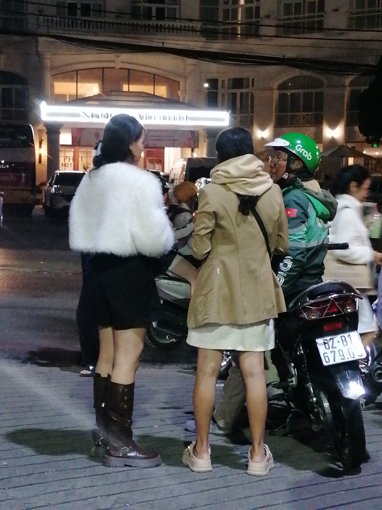 The image captures a street scene at night in Da Lat, Vietnam, likely in front of a hotel named "NGỌC LAN HOTEL" as indicated by illuminated signage. Three individuals are the primary subjects, gathered near a parked motorcycle.
On the left, a woman stands facing away from the camera, wearing a fluffy white jacket over a black outfit and tall brown boots. She appears to be in conversation with another person.
In the center, a person wearing a tan coat and a light-colored skirt is also facing away, engaged in the conversation. Their posture suggests they are standing.
To the right, a person wearing a green "Grab" helmet and a dark jacket with a Vietnamese flag emblem is partially visible. They are also engaged in the interaction.
The setting is a paved area, possibly a sidewalk or street, illuminated by artificial lights. The background shows a building with a well-lit facade and windows. The overall atmosphere appears casual and social, with people interacting outdoors at night. The weather is not discernible from the image. The license plate on the motorcycle reads "62-B1 679.G1".