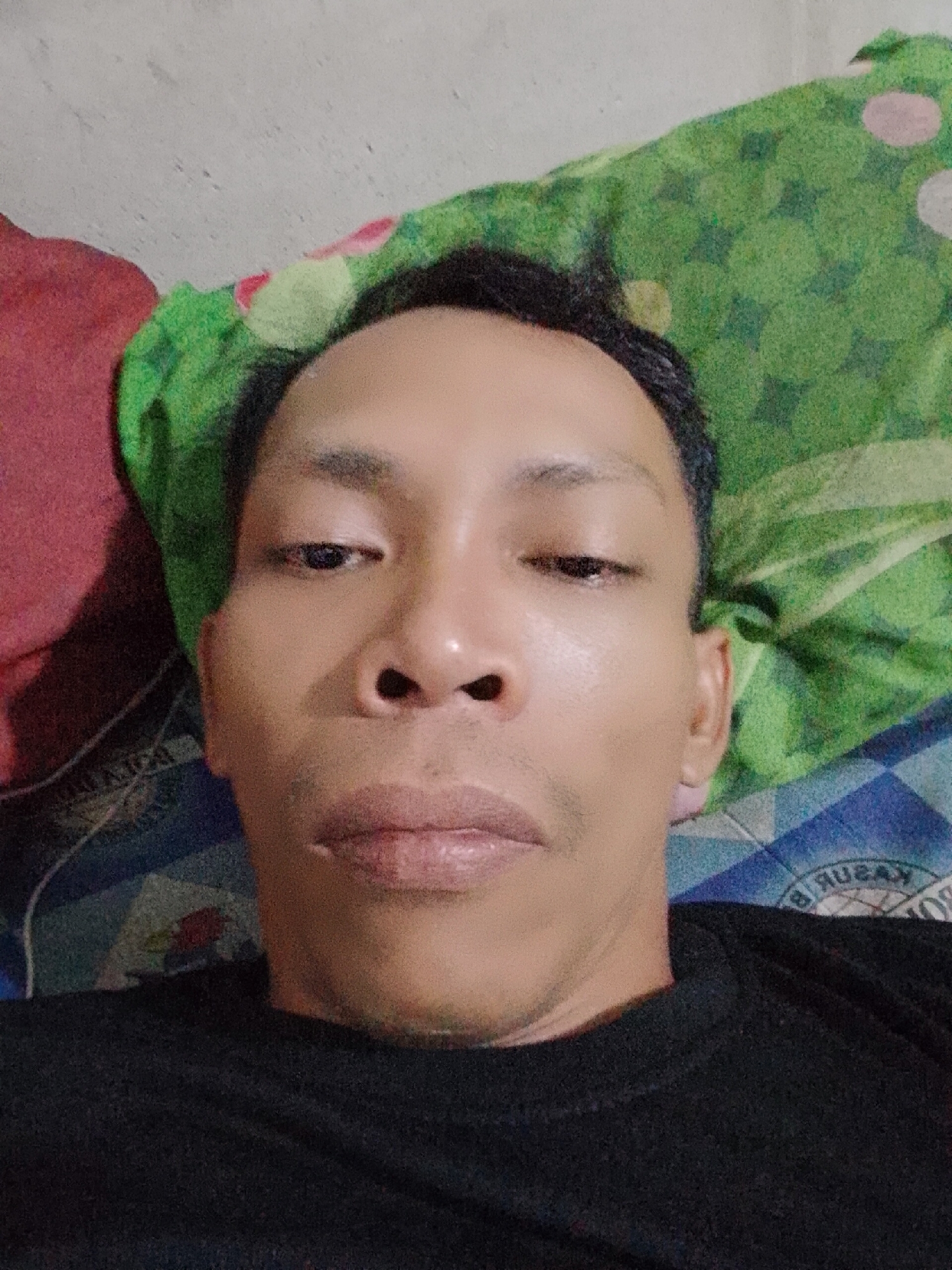 A close-up, top-down perspective photograph shows a man lying on a bed. The man has dark hair and brown skin. He is wearing a black shirt. A green, red, and white patterned bedspread is behind his head. A blue and white patterned sheet is visible below him. The image was taken in Bukit Ketok, Indonesia.