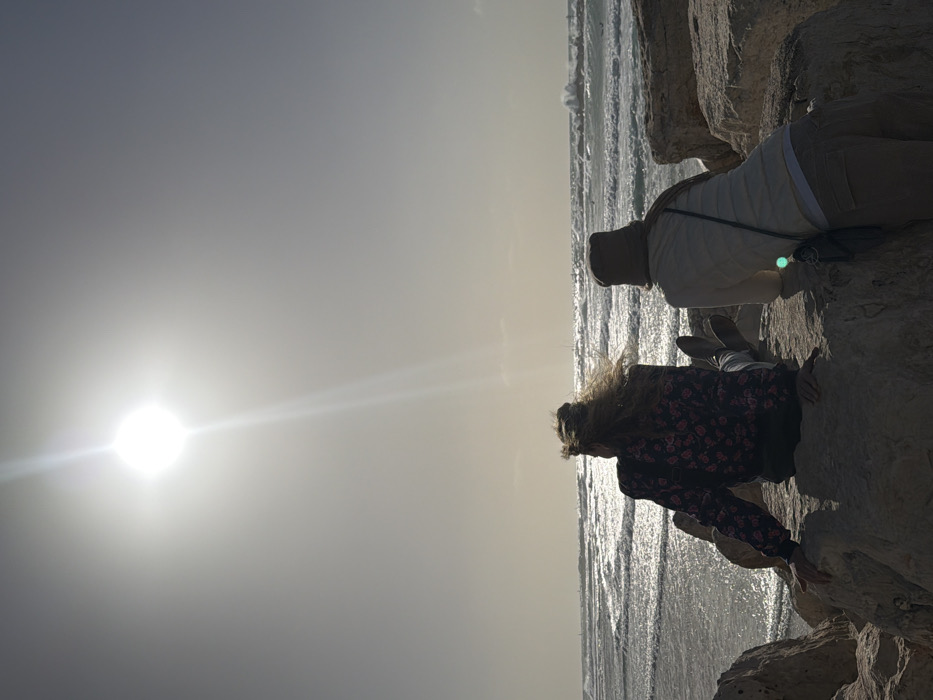 The image depicts a coastal scene in Tel Aviv-Yafo, Israel, with two individuals seated on large, light-colored rocks, facing the Mediterranean Sea. The sun, high in the upper-left, creates a strong glare and backlighting effect, casting the foreground and subjects into partial silhouette.
The individual on the left has light-colored hair styled in a bun, with strands appearing to be windblown. They wear a dark, long-sleeved top with a floral pattern and light-colored trousers, with flat shoes visible. Their right arm rests on a rock. The individual on the right wears a light-colored hat, a scarf, a light-colored long-sleeved top over a lighter undershirt, and light-colored pants. A dark cross-body bag is visible on their right side. Both individuals are observed from behind, looking out at the sea.
The sea exhibits moderate waves, with whitecaps visible across the water. The sun's reflection creates a shimmering effect on the water's surface. A prominent wave is breaking in the mid-ground towards the right, generating a visible splash of white foam. The sky is bright and hazy. Faint structures or vessels are discernible on the distant horizon, partially obscured by atmospheric conditions and glare. The rocks in the foreground are irregularly shaped and appear to form part of a sea wall or breakwater.