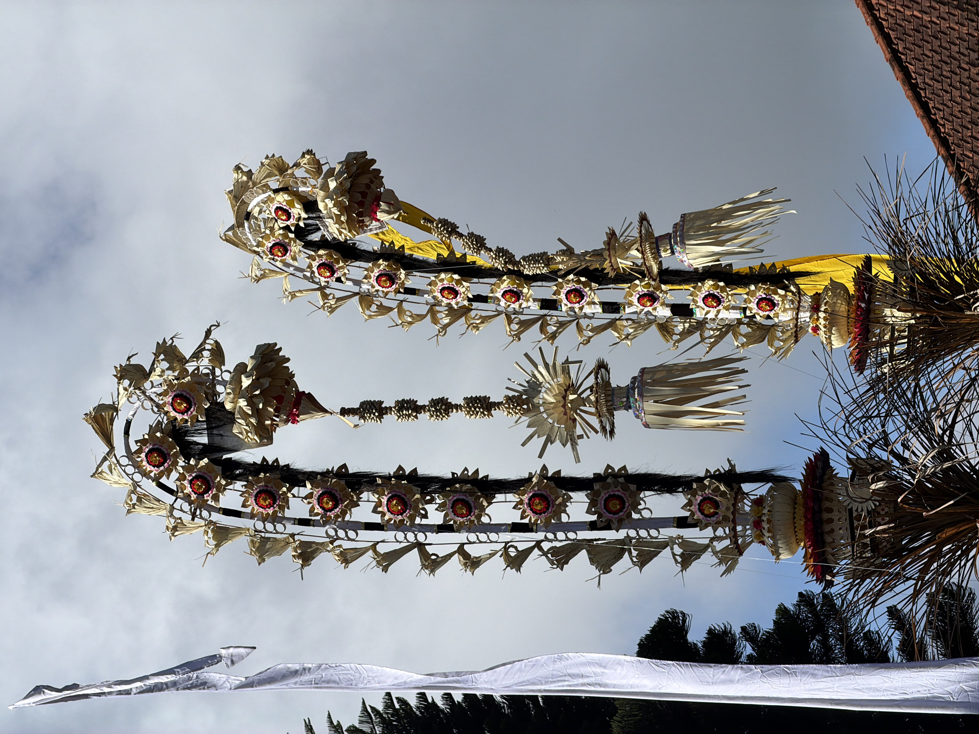 The image, taken in Baturiti, Indonesia, shows two elaborately decorated ceremonial poles against a cloudy sky. A white flag flies on the left, with a portion of a dark green coniferous tree visible behind it. The ceremonial poles are constructed from natural materials, primarily palm leaves, woven and arranged into decorative elements. These elements include tiered bases adorned with red and yellow accents, resembling layered petals or offerings. Suspended from the poles are intricate floral-like ornaments with red and yellow centers, framed by dried palm fronds. Black streamers hang from the upper sections of the poles. Further down, cylindrical, bead-like decorations are strung along one of the poles, and a large, starburst-shaped ornament made of woven palm leaves is visible on the right pole. The background also shows a glimpse of a red tiled roof on the lower right. The overall impression is one of festive or religious decoration, likely for a cultural event or ceremony.