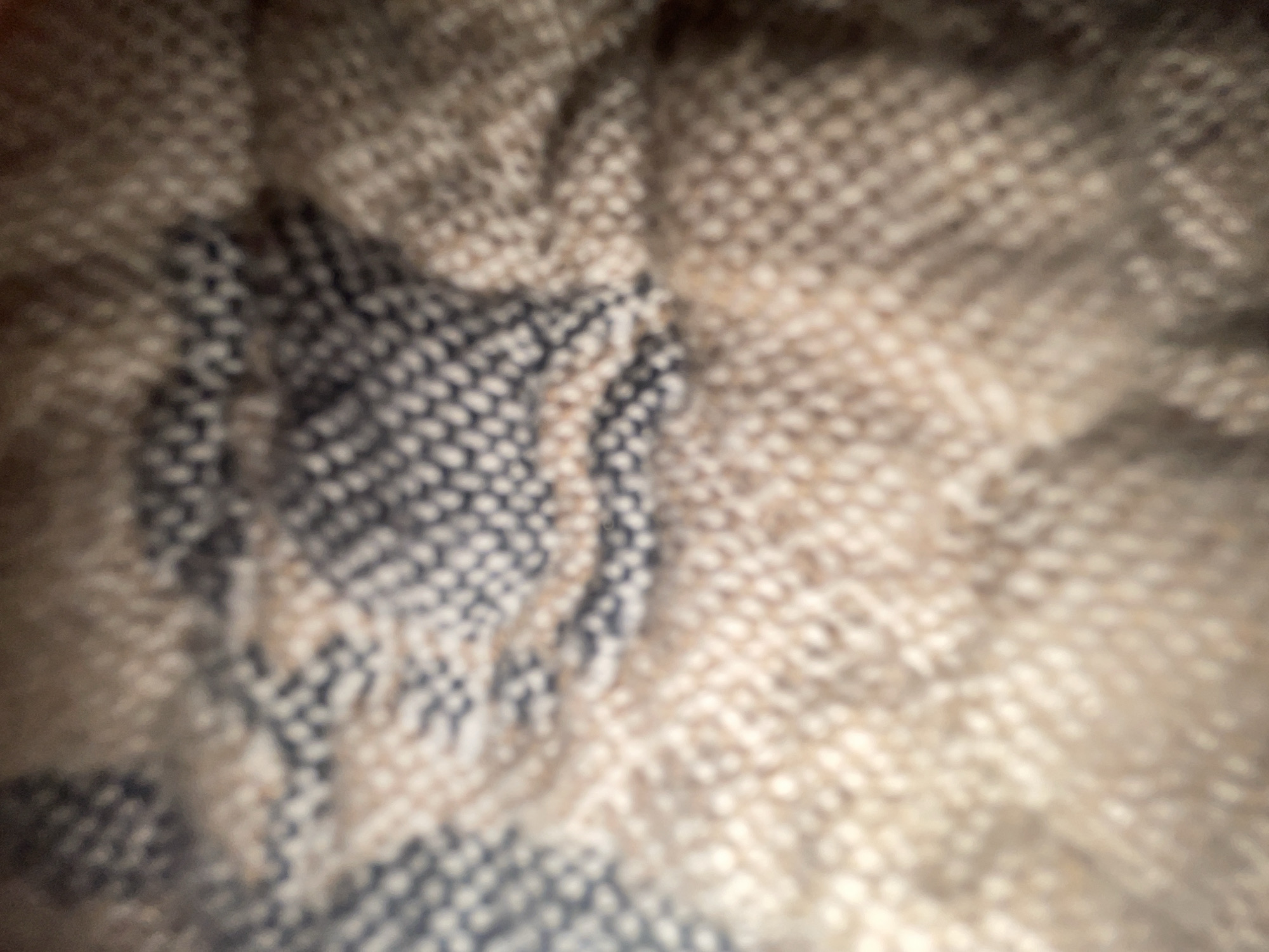 The image is a close-up, out-of-focus shot of a textured fabric, possibly a blanket or a rug. The fabric has a woven pattern with alternating sections of dark blue and lighter beige or tan threads. The texture appears to be coarse, with a visible weave structure even in the blur. The lighting is dim and diffused, casting soft shadows across the fabric. There are no people, objects, or environmental details that could place this image in Vladikavkaz, Russia, or any other specific location. The focus is entirely on the material and its texture.