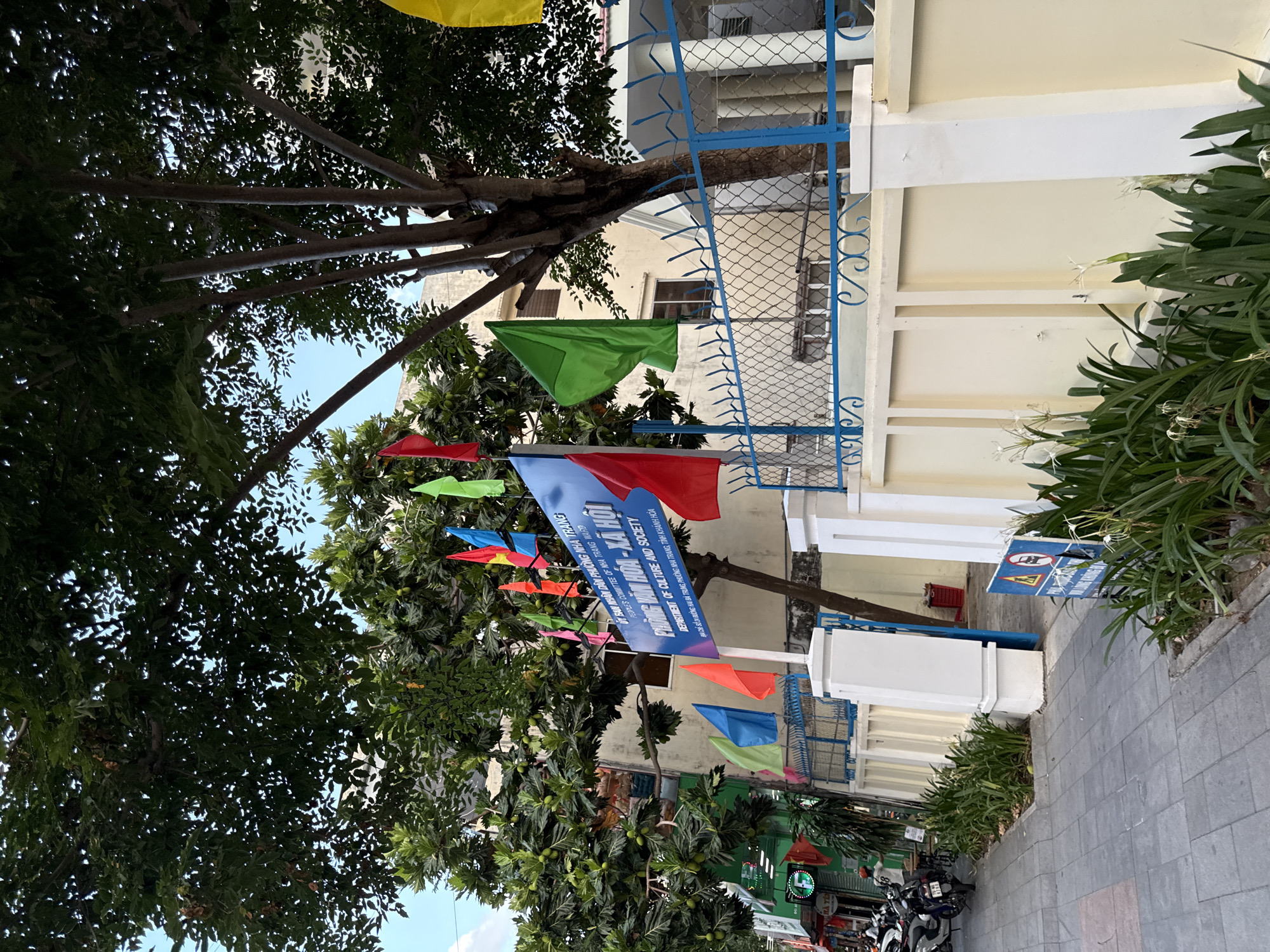 The image shows a street in Nha Trang, Vietnam, with a building displaying a blue sign that reads "PHÒNG VĂN HÓA - XÃ HỘI" and "DEPARTMENT OF CULTURE AND SOCIETY". Numerous colorful flags, including red, blue, green, and yellow, are displayed in front of the building. A sidewalk runs alongside the street, with several motorcycles parked on the left. A fence with blue accents surrounds the property. Lush green plants, including flowers, are visible in front of the wall. The sky is blue with some clouds. The overall scene depicts an outdoor public space, likely a government office or community center.